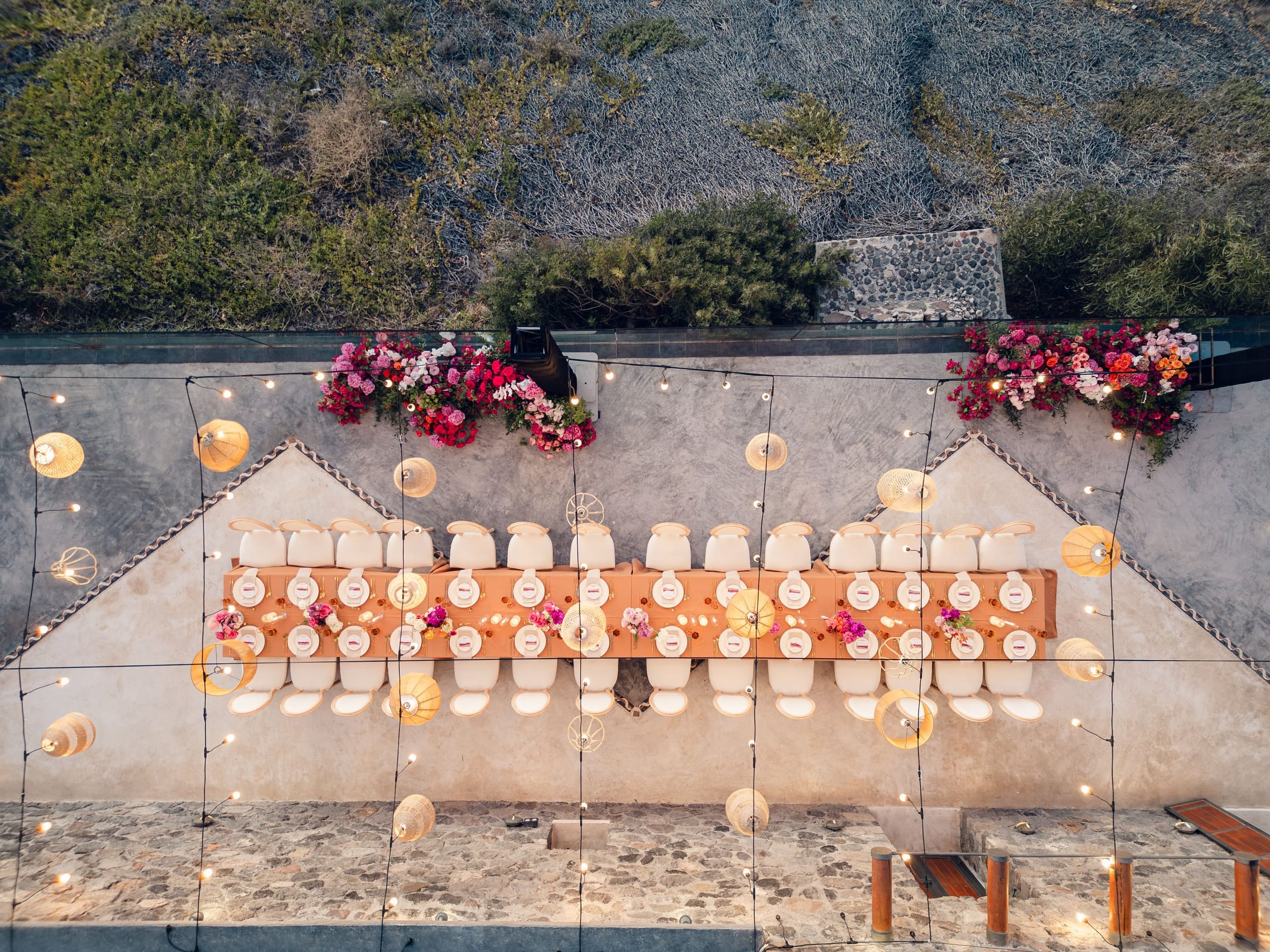 An overhead aerial view of a long wedding dinner table on a stone terrace at Cavo Ventus Luxury Villa in Santorini, Greece. A runner of warm terracotta linen runs down the centre, lined with white chairs, white plates and small posies of pink and magenta blooms, with bougainvillea and roses spilling along the stone wall at each end. Strings of warm lights and woven rattan lanterns hang overhead above the guests' places.