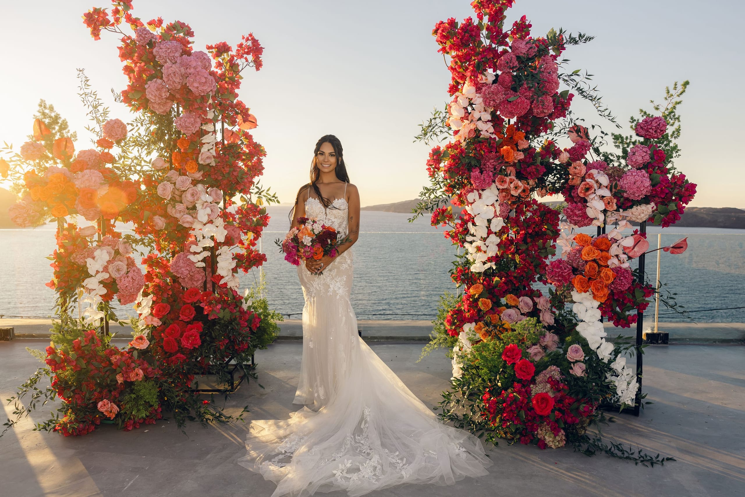 A bride in a fitted white lace gown with a long train stands smiling between two tall floral arches on a terrace at Cavo Ventus Luxury Villa in Santorini, Greece. The arches are densely packed with pink and red bougainvillea, pink and orange roses, white orchids, and orange and coral blooms, with olive foliage trailing at their bases. She holds a bouquet of orange, magenta and deep red flowers, and behind her the calm sea and distant hills glow in warm golden sunset light.