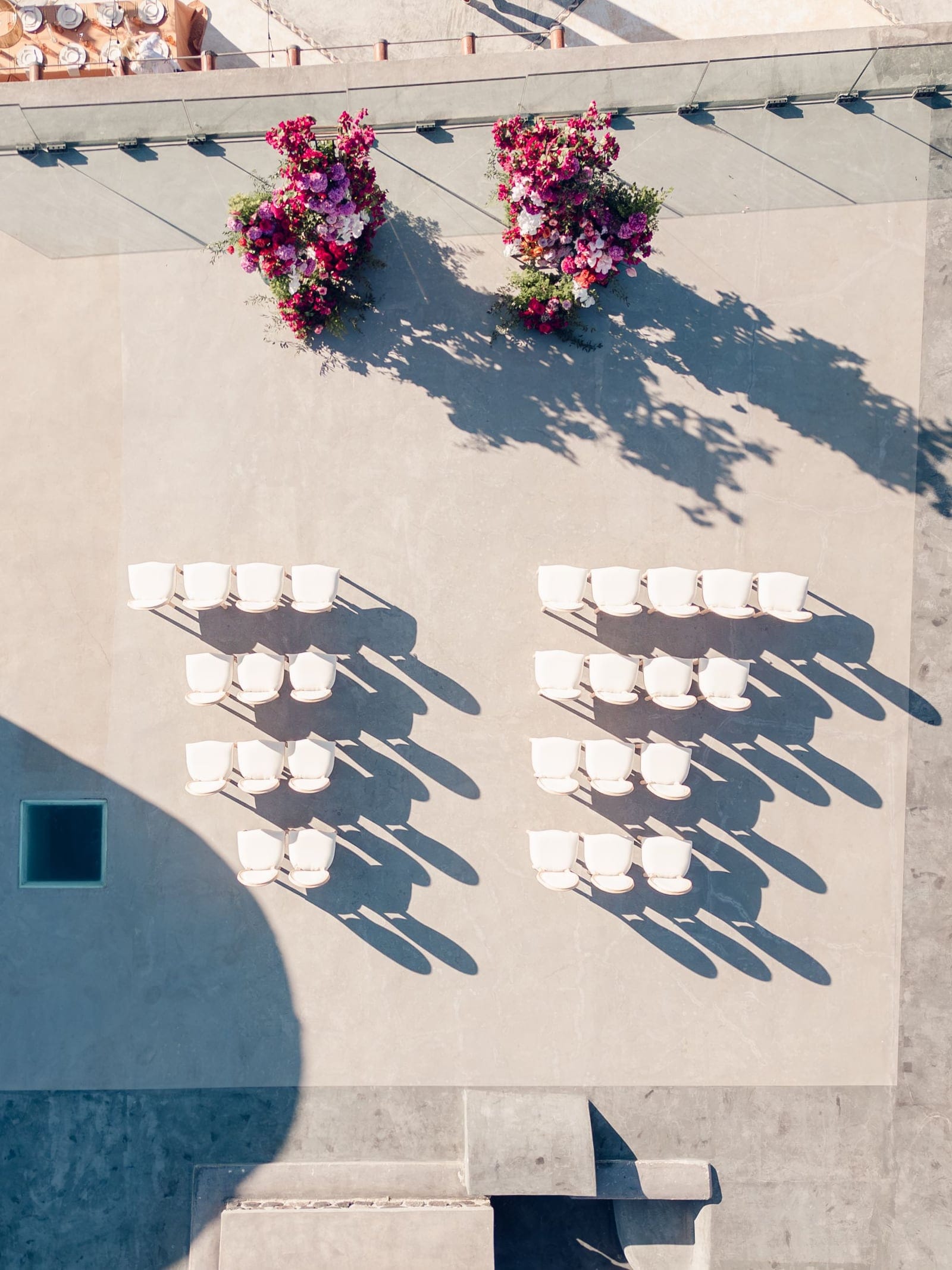 Seen from above, two symmetrical blocks of white cushioned chairs face an open pale stone terrace at Cavo Ventus Luxury Villa in Santorini, Greece, arranged for a wedding ceremony. At the top of the terrace stand two tall floral arrangements of bougainvillea in magenta, deep pink and violet, with touches of white blooms and green foliage. Strong afternoon sun casts long, sharp shadows of the chairs and flowers across the stone, creating a calm, sunlit mood.