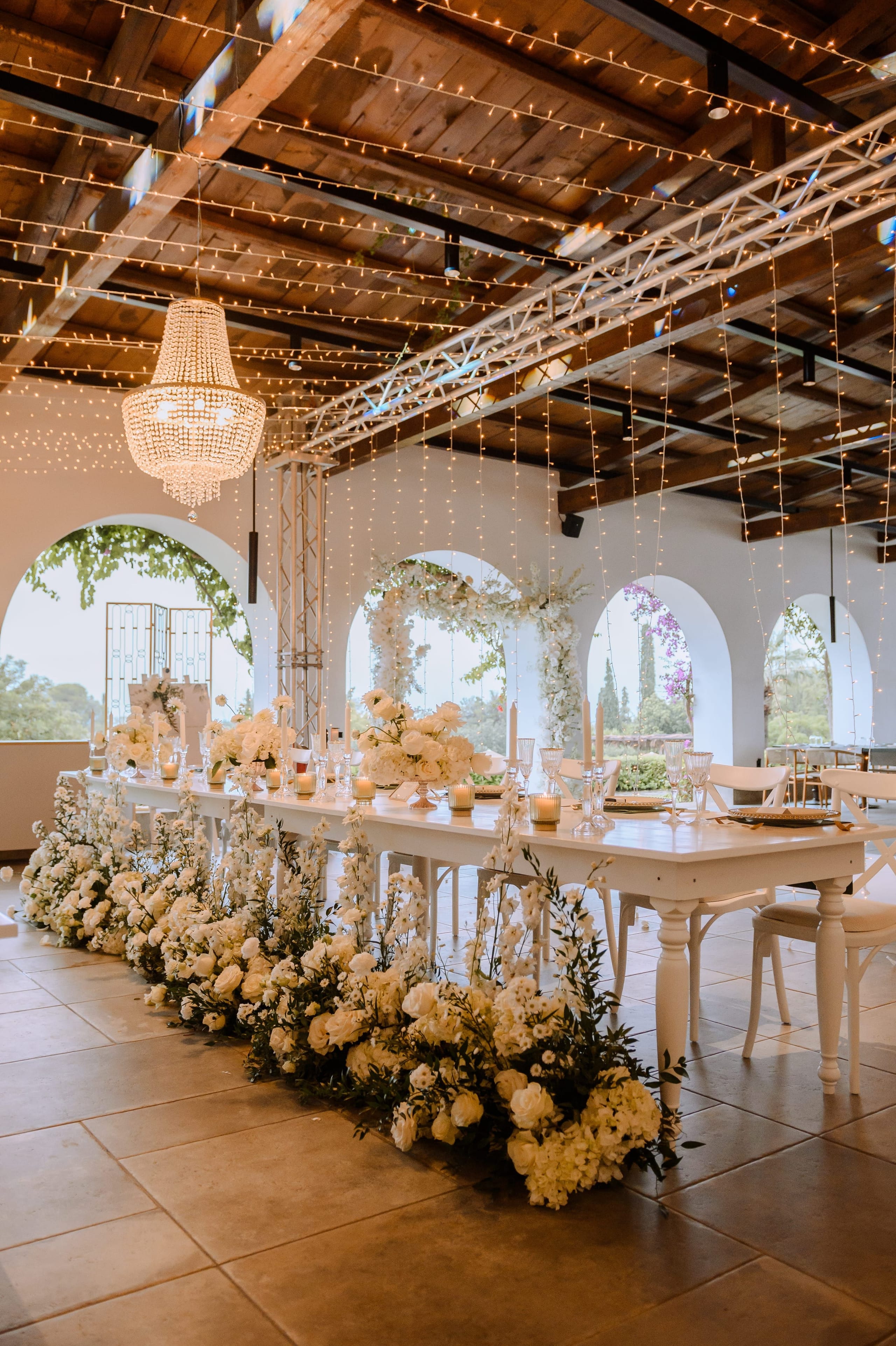A long white banquet table runs through a rustic, string-lit hall at Minos Beach Art Hotel in Agios Nikolaos, Crete, Greece, its base buried under a lush, cascading garland of white roses, hydrangeas and sprigs of olive foliage. Tall candles, glassware and a central arrangement of white blooms sit along the table, which is set with cross-back chairs. Arched white openings frame green views beyond, and a crystal chandelier hangs above, creating a soft, romantic wedding table scene.
