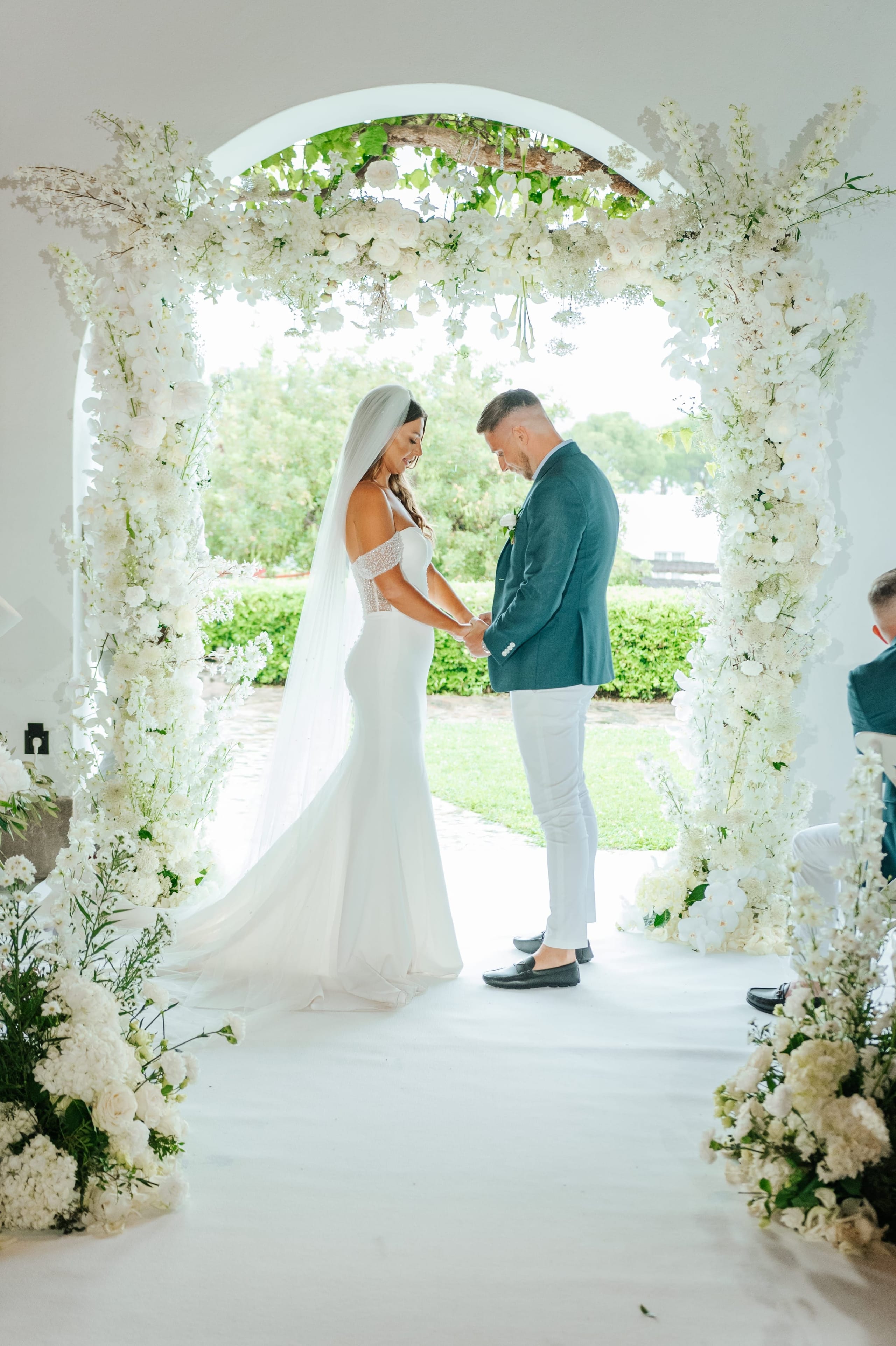 A bride in a fitted white gown with a long veil and a groom in a teal jacket and white trousers stand hand in hand, smiling down, beneath a white wedding arch dense with roses, delphinium spires and hydrangeas. The arch frames a green lawn and trees beyond the white walls, and bright, soft light falls across the scene. This is a wedding ceremony flower arch at Minos Beach Art Hotel in Agios Nikolaos, Crete, Greece.