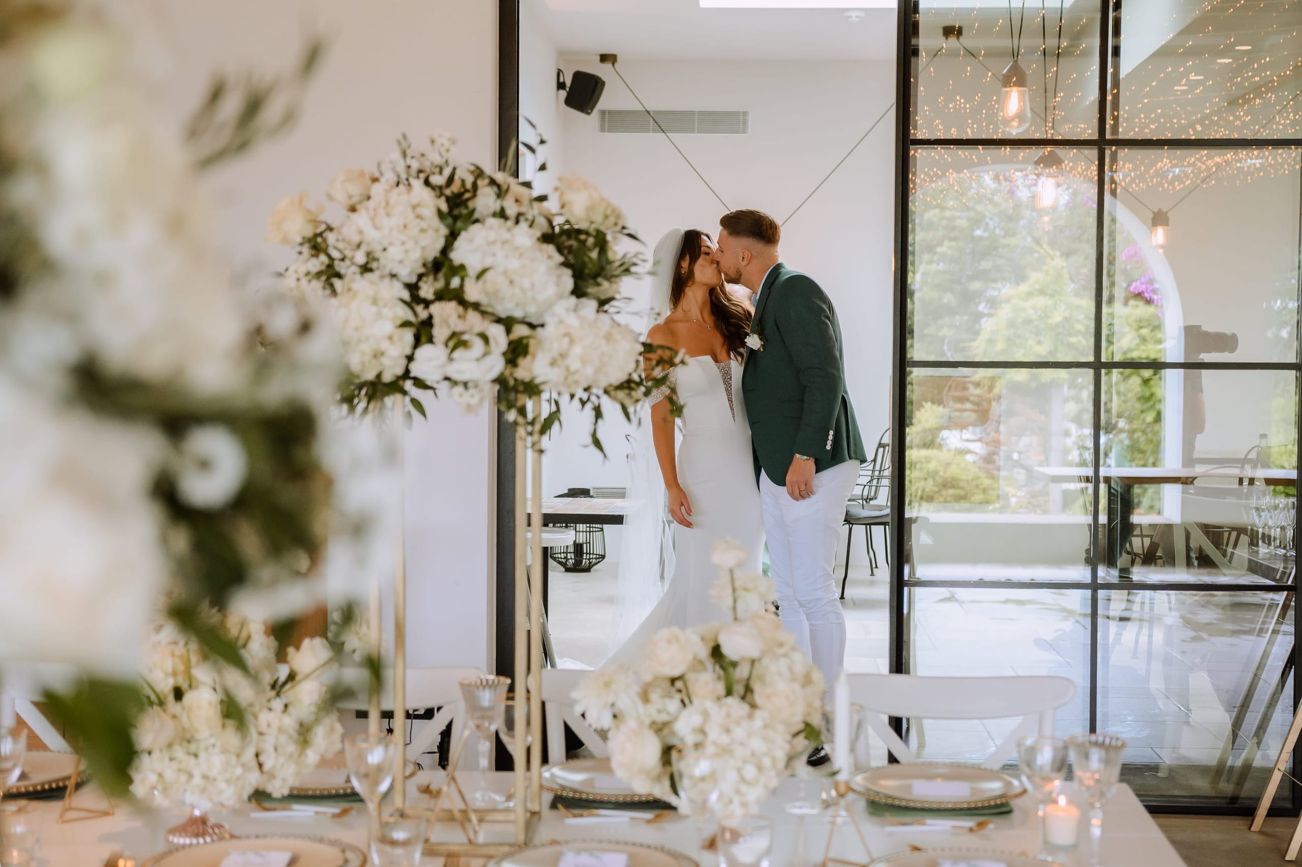 A bride in a fitted white gown and a groom in a dark green jacket and white trousers share a kiss in a bright white room, framed by tall glass doors. In the foreground, a wedding table in Greece is dressed with white hydrangeas and garden roses on gold stands, with candles and gold-rimmed plates. Through the doors, string lights and green shrubs hint at the terrace at Minos Beach Art Hotel in Agios Nikolaos, Crete.