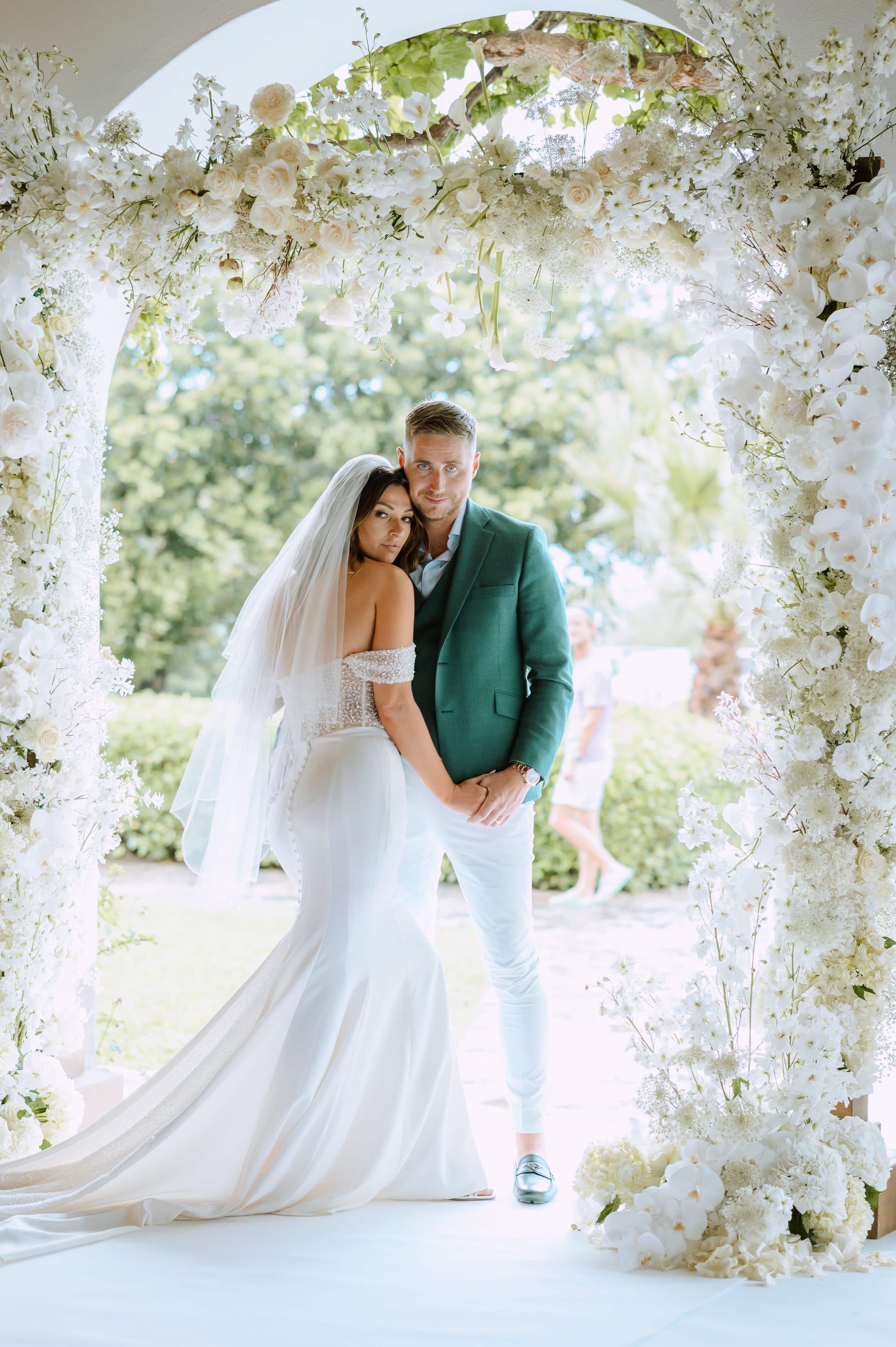 A bride and groom stand together under a tall white floral arch, the bride's head resting on the groom's shoulder as they look toward the camera. The arch is covered in white garden roses, orchids, and delicate sprays of baby's breath, and the couple is framed by green trees in soft focus at the wedding at Minos Beach Art Hotel in Greece. The bride wears a veil and an off-the-shoulder beaded white gown, while the groom wears a green suit and light trousers.