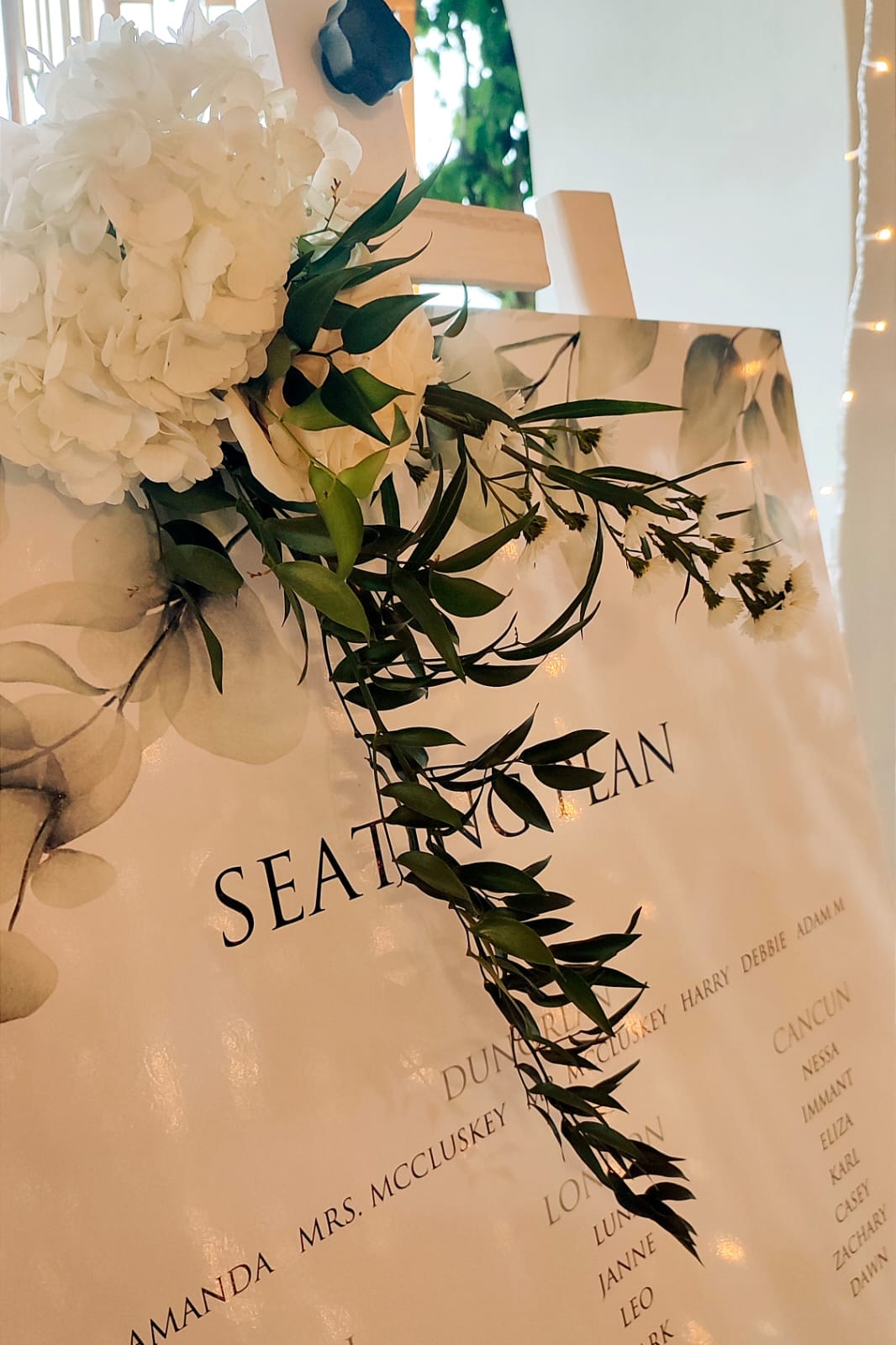 A wedding seating plan on an easel, its cream board printed with faint grey eucalyptus leaves and black serif lettering. Cascading down the board is a trailing spray of slender green foliage with tiny white blooms, and at the top sits a large cluster of white hydrangea beside cream roses. This wedding table display is at Minos Beach Art Hotel in Agios Nikolaos, Crete, Greece, with soft string lights glowing at the edge of the frame.