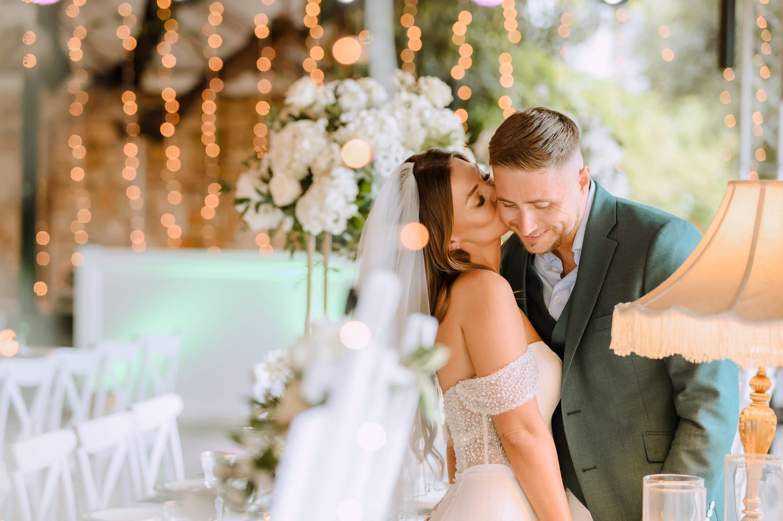 A bride in an off-the-shoulder gown with a pearl-beaded bodice kisses the cheek of a smiling groom in a green suit, their eyes closed in a tender moment. Behind them, tall white hydrangea arrangements rise above the table, with more white blooms in the foreground, all framed by strings of warm fairy lights at Minos Beach Art Hotel in Agios Nikolaos, Crete, Greece. The soft bokeh and golden lamplight give the wedding reception a romantic, intimate glow.