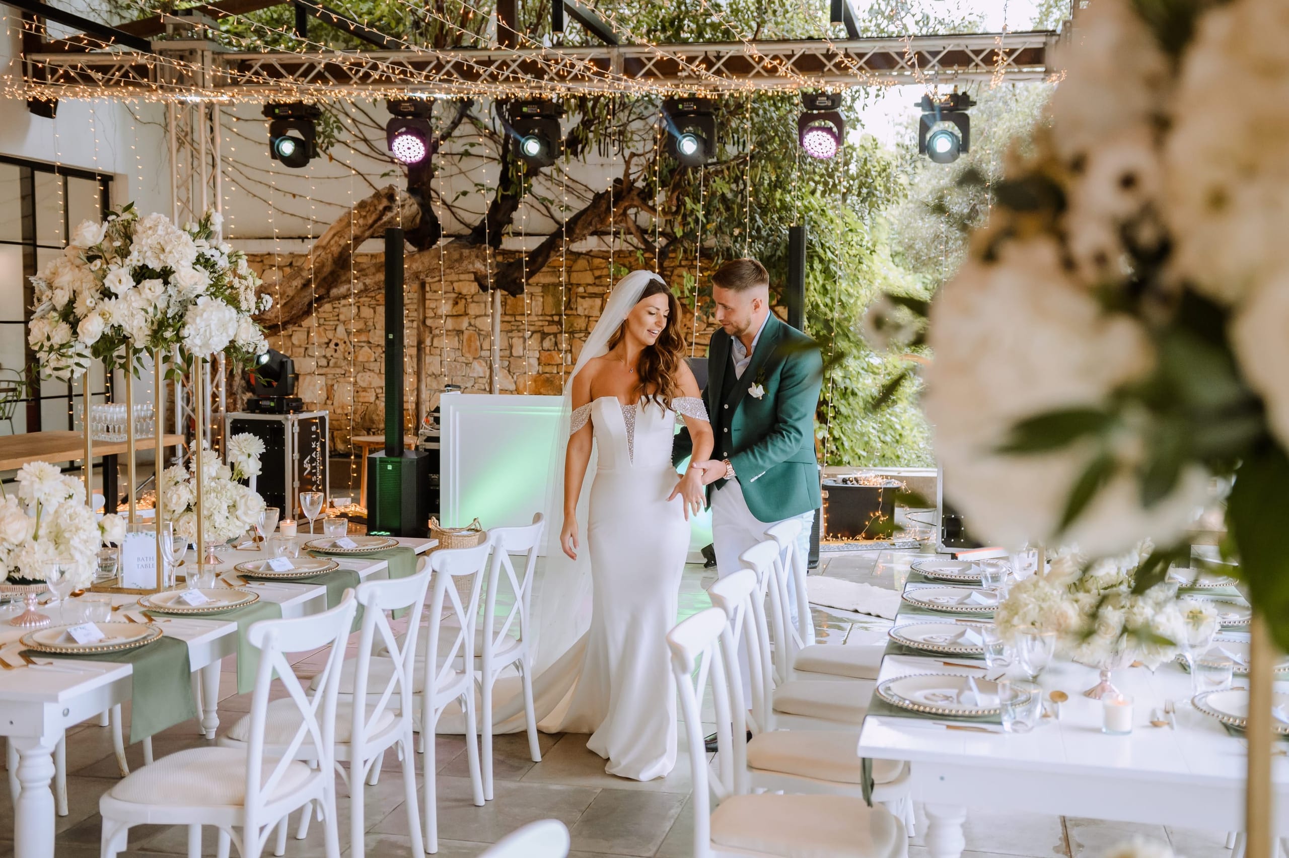 A bride in a fitted white off-shoulder gown with a long veil and a groom in a deep green suit stroll arm in arm between long tables at a wedding reception in Crete, Greece, at Minos Beach Art Hotel. Tall gold stands hold lush white hydrangeas and roses, the same blooms repeated in low arrangements down the tables, and the place settings sit on sage-green runners. Strings of fairy lights hang beneath a rustic beam structure, with an old stone wall and green foliage beyond.