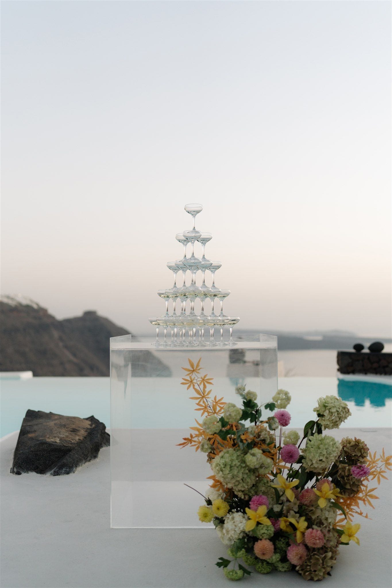 A pyramid of champagne coupe glasses rises on a clear acrylic plinth beside an infinity pool at Aenaon Villas in Santorini, Greece, with the caldera and pale dusk sky stretching behind it. At the base sits a loose arrangement of green hydrangeas, pink dahlias, yellow lilies and trailing orange-tipped foliage, a wedding display with a soft, modern feel. The white terrace, the dark volcanic rock and the muted pink-grey light make the scene calm and airy.