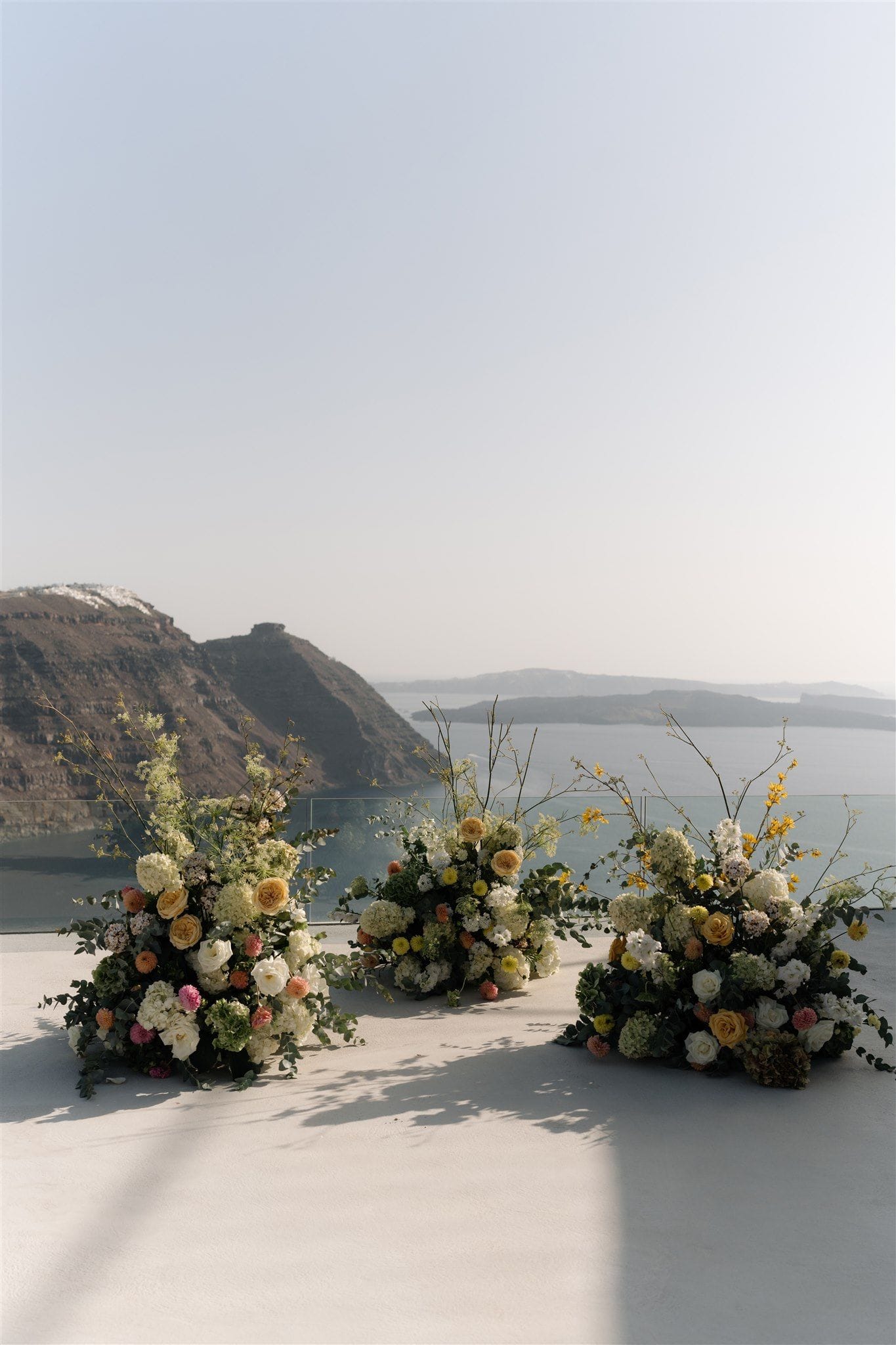 Three low, lush floral installations sit on a pale stone terrace at Aenaon Villas in Santorini, Greece, their rounded shapes spilling with cream hydrangeas, white and apricot garden roses, pink and orange dahlias, and trailing eucalyptus, with slender yellow-flowered branches rising above them. Beyond the terrace edge, the caldera's dark volcanic cliffs and the hazy blue sea stretch under a soft, pale sky. These wedding flowers are arranged for a ceremony, with the cool, sun-washed setting giving the scene a calm, airy feel.