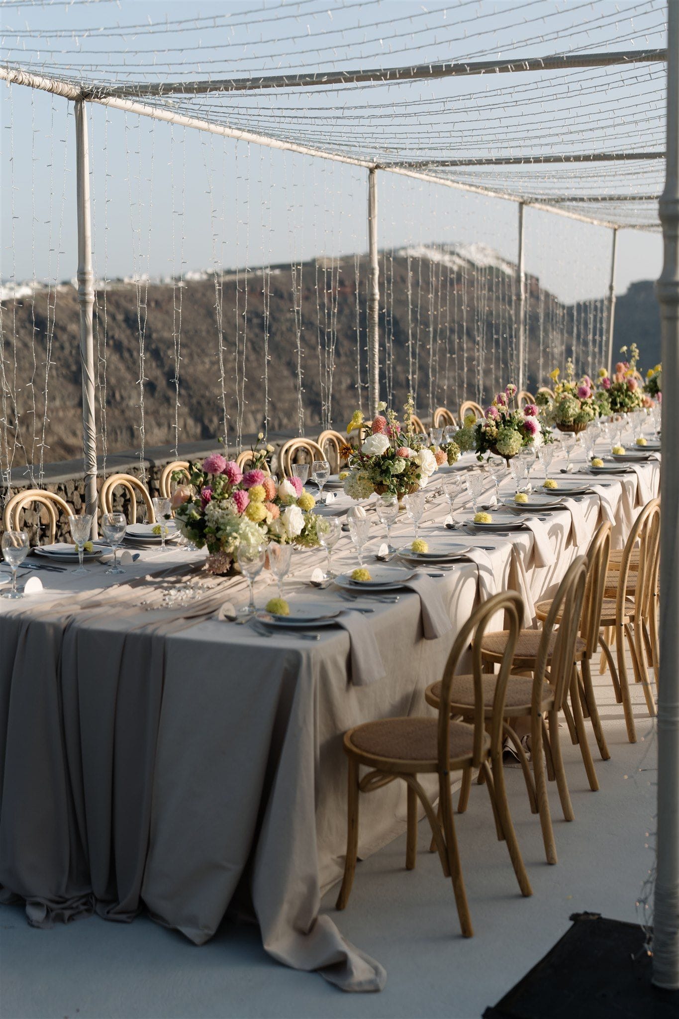 A long wedding dinner table draped in soft grey linen runs along a terrace at Aenaon Villas in Santorini, Greece, set with clear glassware, pale plates and low centrepieces of pink, cream and yellow blooms, including what look like garden roses and hydrangeas. Bentwood chairs line both sides beneath a canopy of fine string lights, with a dramatic volcanic cliff and whitewashed village edge in the soft, late-afternoon light beyond.