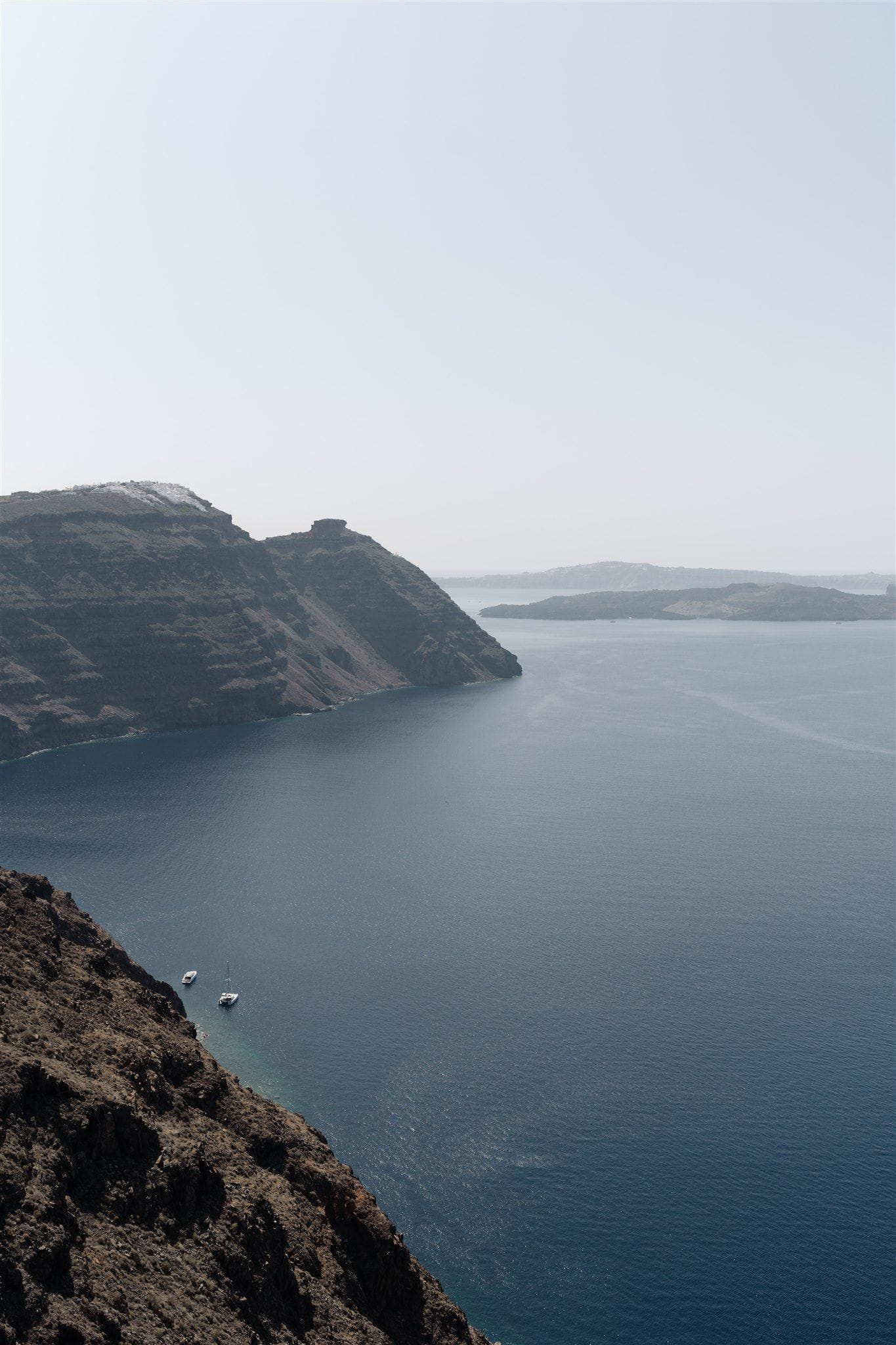 Looking down from a rugged volcanic cliff at Aenaon Villas in Santorini, Greece, across a wide, deep-blue caldera sea under a pale, hazy sky. Dark brown rock layers rise on the left, and two small white boats sit at anchor near the base of the cliff, while faint islands fade into the distance on the right. It is a quiet, expansive view, the kind of backdrop chosen for a wedding story.