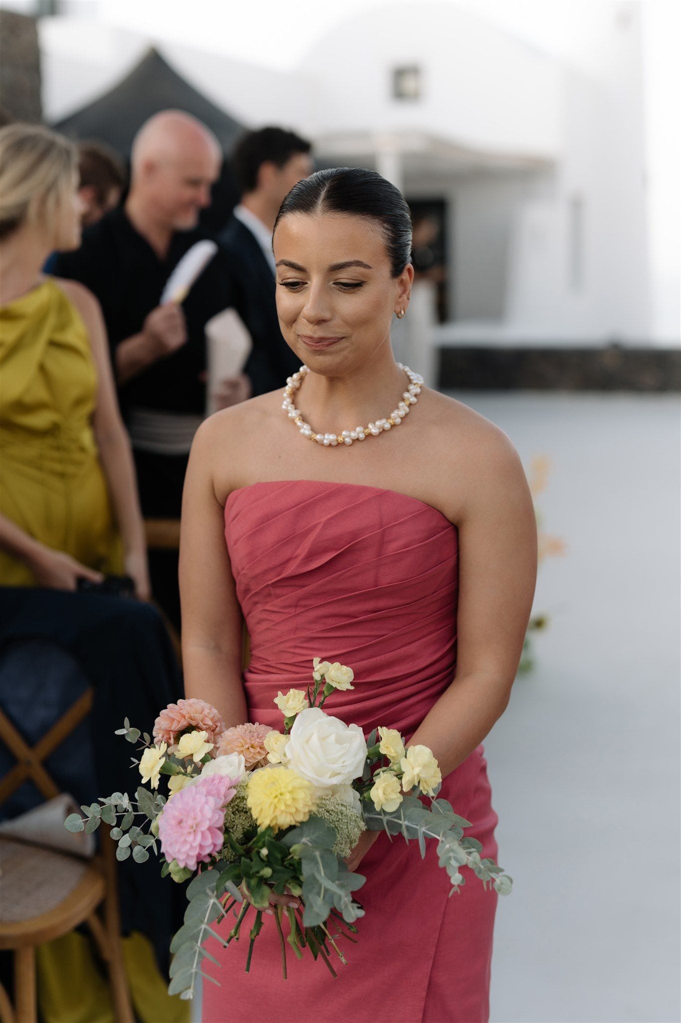 A bridesmaid in a strapless rose-pink gown with soft pleated detail stands on a white terrace, holding a loose hand-tied bouquet of pale yellow roses, blush dahlias, a white garden rose, and feathery green seed heads wrapped in silvery eucalyptus. She wears a pearl necklace and gazes down at the flowers with a gentle smile, while guests in mustard and black sit softly blurred behind her. This wedding ceremony took place at Aenaon Villas in Santorini, Greece, with whitewashed Cycladic buildings in the background.