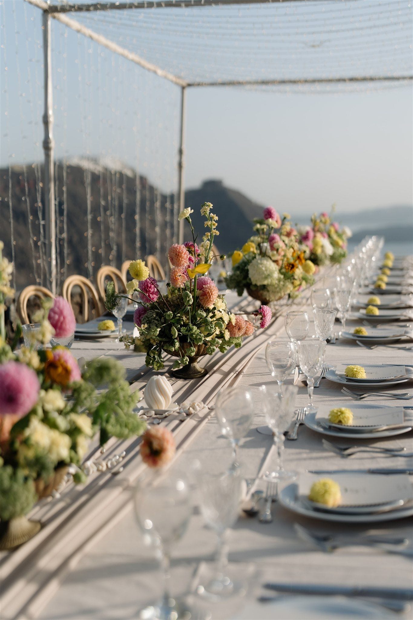 A long wedding table on a clifftop terrace at Aenaon Villas in Santorini, Greece, set with white linen and clear stemmed glasses. Low centrepieces of pink and peach dahlias, pale yellow blooms and cream hydrangea-like heads sit in brass urns along the table, and small yellow pompom flowers rest on each place setting. Strings of tiny lights hang overhead beneath a pale sky, with the hazy island hills and sea softly blurred behind.