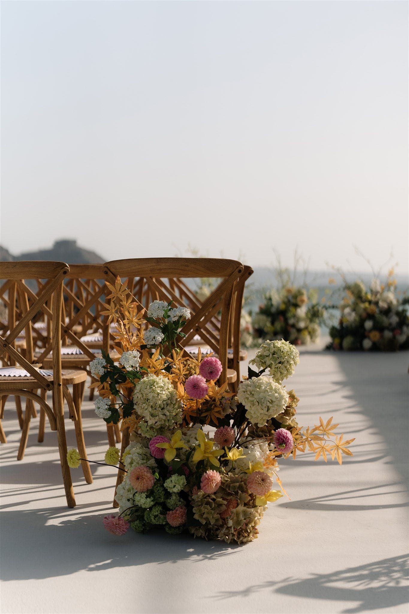 A low, generous arrangement of cream hydrangeas, pink dahlias, yellow lilies and dried golden foliage sits on a pale stone floor, flanking the aisle of wooden cross-back chairs at a wedding ceremony at Aenaon Villas in Santorini, Greece. Soft, late-afternoon sunlight casts long shadows across the terrace, and the sea and a distant headland fade into a hazy sky behind more flower arrangements. The mood is airy, warm and relaxed.