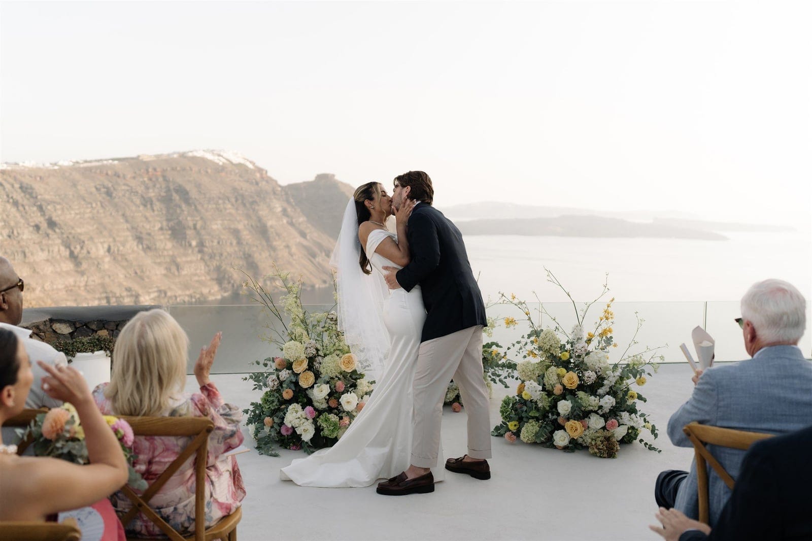 A bride in a fitted white satin gown and long veil kisses her groom in a navy blazer and cream trousers on a white terrace at Aenaon Villas in Santorini, Greece, while guests applaud from wooden chairs. Two large wedding ceremony arrangements of cream and blush roses, white hydrangeas, green foliage and sprays of yellow blooms frame the couple. Behind them, a hazy pale sky meets the sea and the rugged caldera cliffs of Santorini.