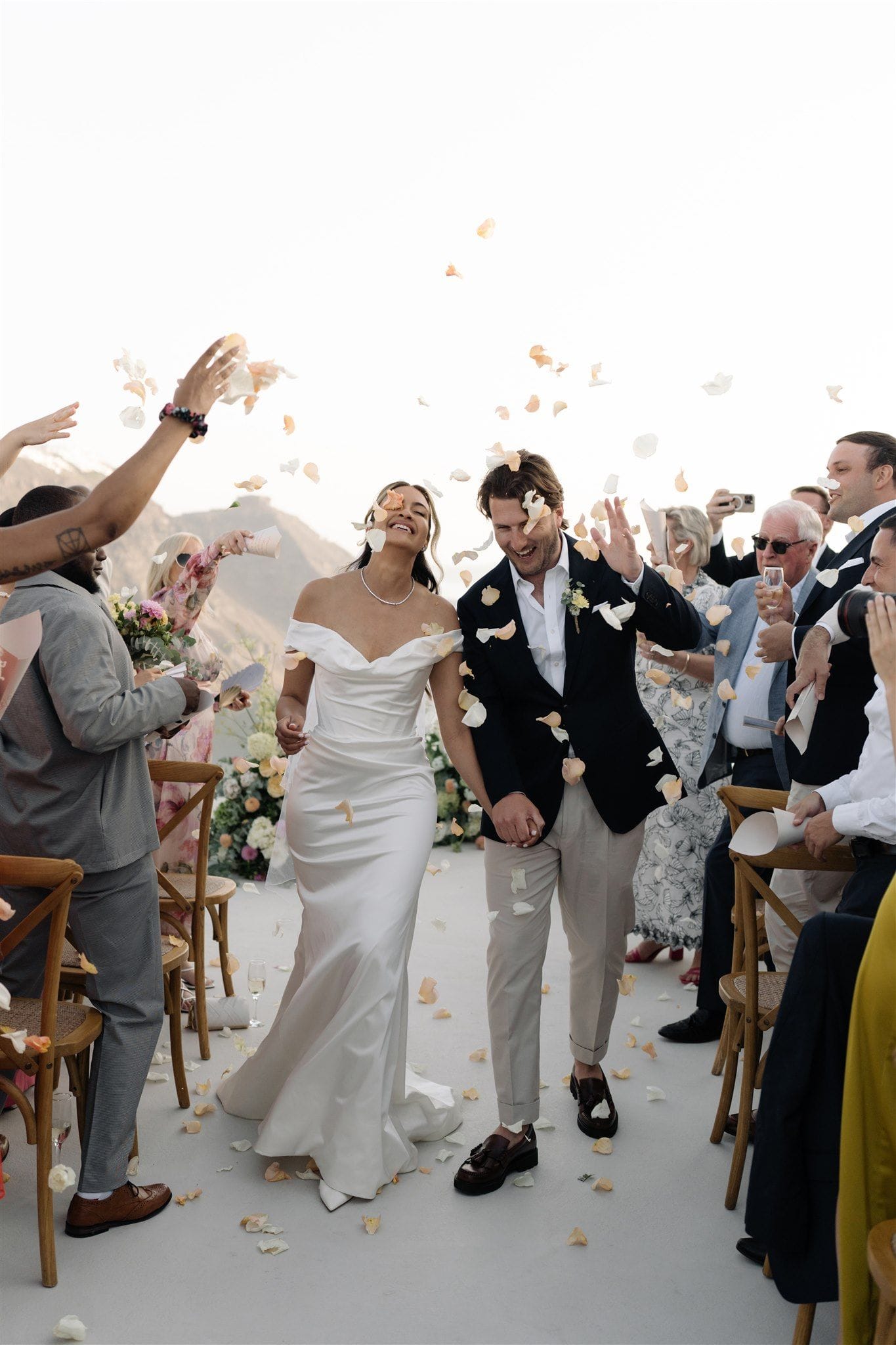 A bride in a white off-the-shoulder satin gown and a groom in a navy blazer and cream trousers walk hand in hand down an aisle, laughing as guests on both sides throw pale peach and white rose petals into the air. Behind them stands a white floral arrangement of hydrangeas and roses, with hazy hillsides and a bright sky beyond, at Aenaon Villas wedding ceremony in Santorini, Greece.