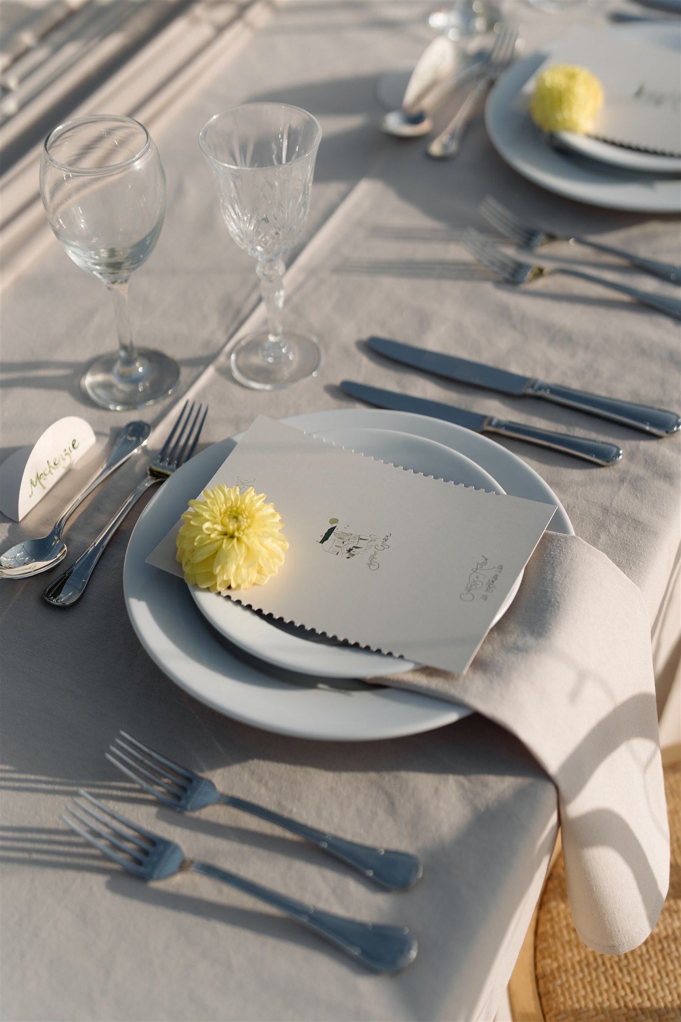 A wedding table setting in Greece, laid on a pale linen tablecloth in warm evening light, with a single yellow chrysanthemum resting on a deckle-edged menu card that sits on stacked white plates. A linen napkin falls beside the plate, and crystal wine glasses, silverware and a small heart-shaped name card complete the table at Aenaon Villas in Santorini. The soft shadows and muted tones give the scene a calm, elegant feel.