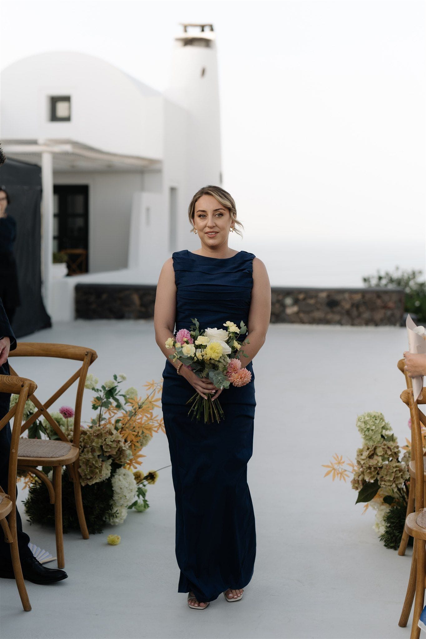 A bridesmaid in a navy, sleeveless floor-length gown stands centred on a white stone aisle at Aenaon Villas in Santorini, Greece, holding a loose wedding bouquet of pink and peach dahlias, pale yellow roses and eucalyptus. Behind her, whitewashed walls and a cylindrical chimney sit against a pale, misty sky, and wooden cross-back chairs line the aisle, dressed with trailing hydrangeas and golden grasses.