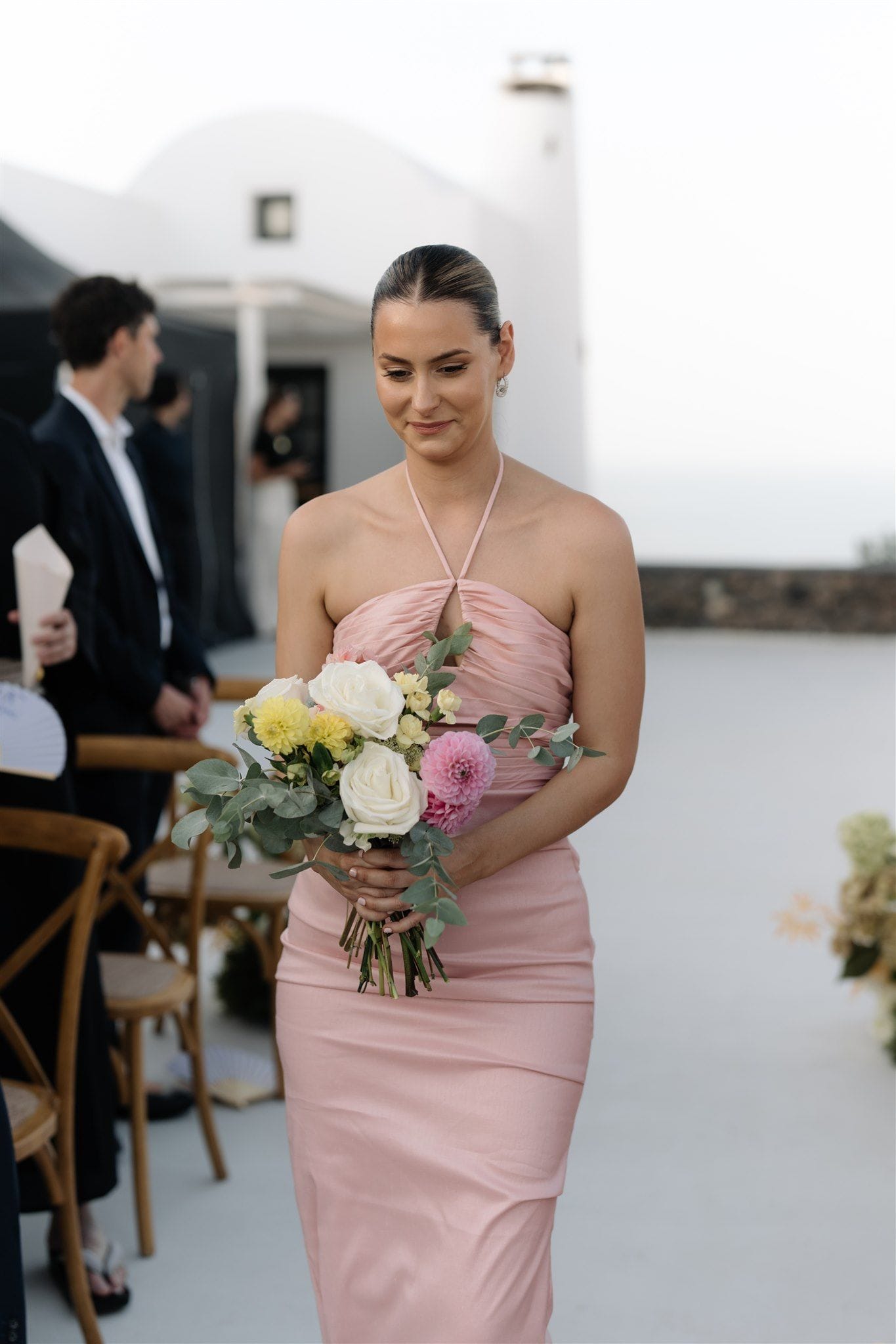 A bridesmaid in a blush pink halter-neck satin gown walks down a white aisle at a wedding ceremony at Aenaon Villas in Santorini, Greece, holding a hand-tied bouquet of white garden roses, yellow dahlias, a pink dahlia and sprigs of eucalyptus. Whitewashed walls and a rounded chimney rise behind her, and wooden chairs line the aisle where guests sit in dark clothing, with more cream and green floral arrangements along the edge of the path.