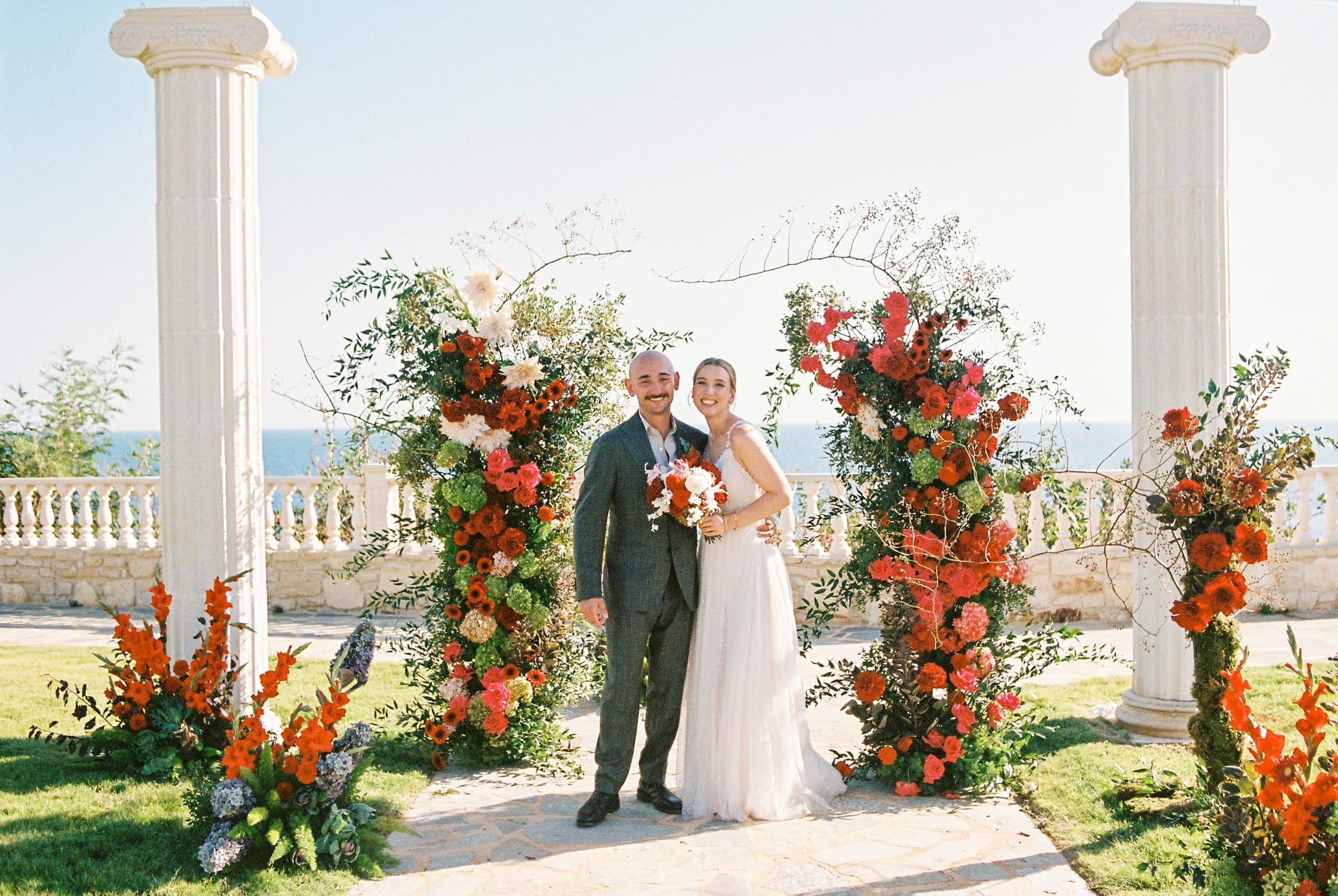 A bride and groom stand smiling between two tall floral arches at a wedding at Wine Dark Villas in Greece, framed by white Doric columns. The arches are packed with red and coral garden roses, orange ranunculus, pink hydrangeas, pale dahlias and sprays of green foliage. The groom wears a grey suit and the bride a flowing white gown, holding a bouquet of red and cream blooms, with the sea glimmering behind a stone balustrade under a clear sky.