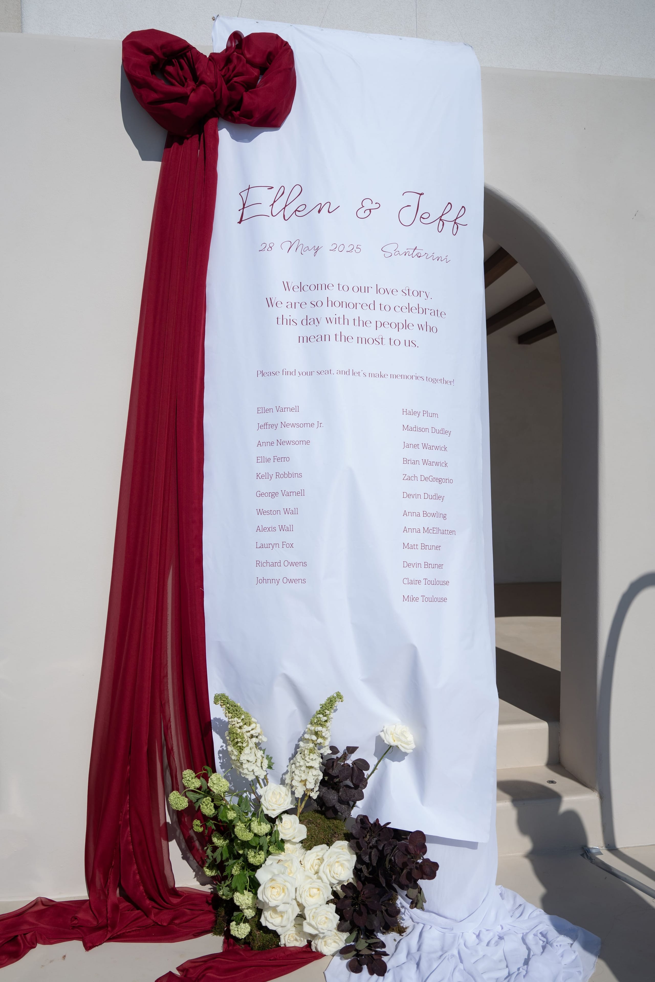 A tall white seating chart banner hangs against a whitewashed wall at Islet Santorini in Thira, Greece, its top gathered by a deep burgundy chiffon bow with long red streamers trailing down. Printed in burgundy script are the names Ellen and Jeff and their wedding date, with a welcome message above a list of guest names. At the base, white roses, cream-green flowers and dark plum foliage spill over the fabric, with a shadowed archway to the right.