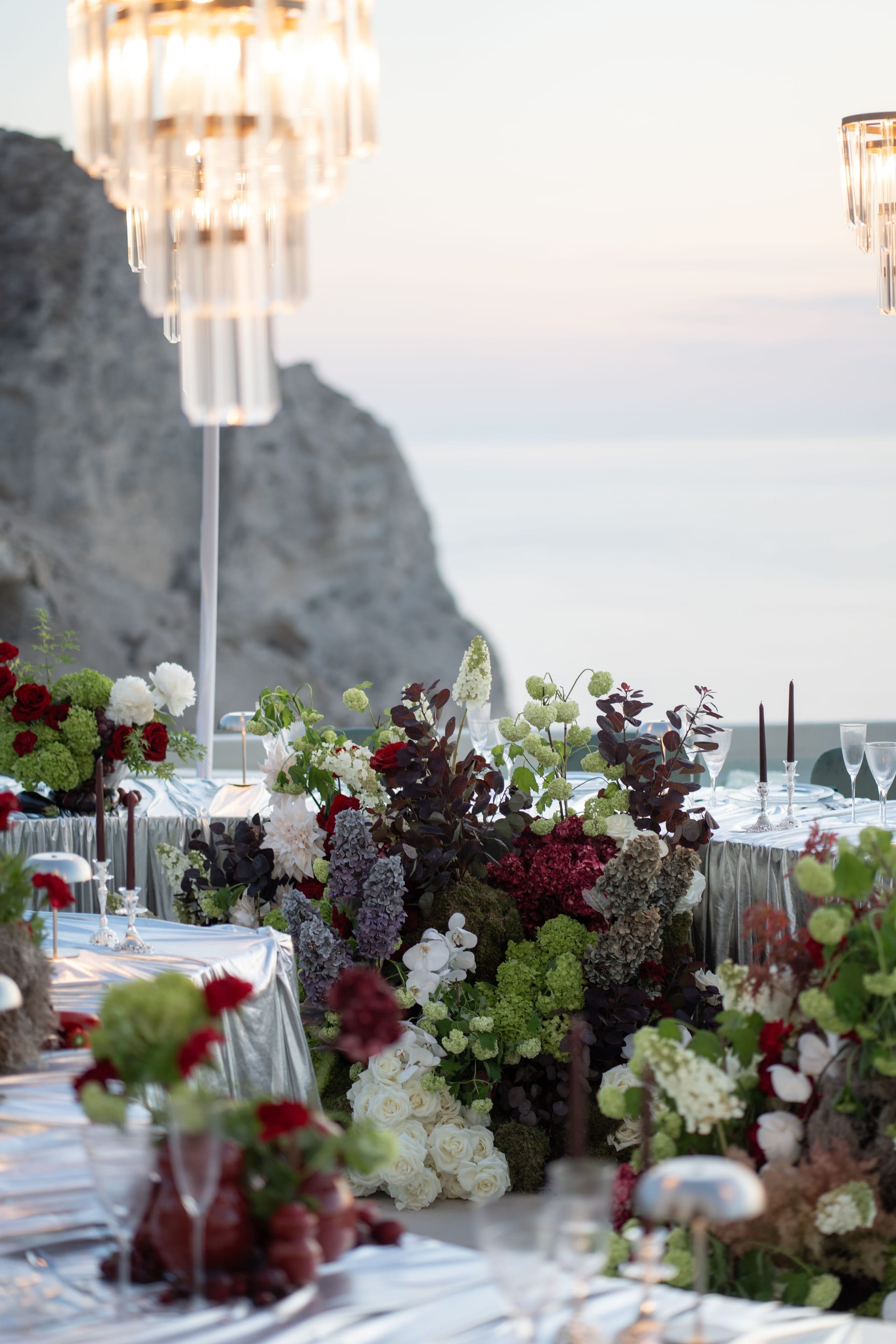 A long wedding table at Islet Santorini in Thira, Greece, dressed in silver-grey linen and set for dinner at dusk, with a sheer, pale sea behind it. Down the centre runs a lush low arrangement of red garden roses, white roses, deep burgundy blooms, green hydrangea, white dahlias and dark plum foliage, with lilac-toned stems rising in between. Tall crystal chandeliers hang above, and a pale cliff rises on the left.