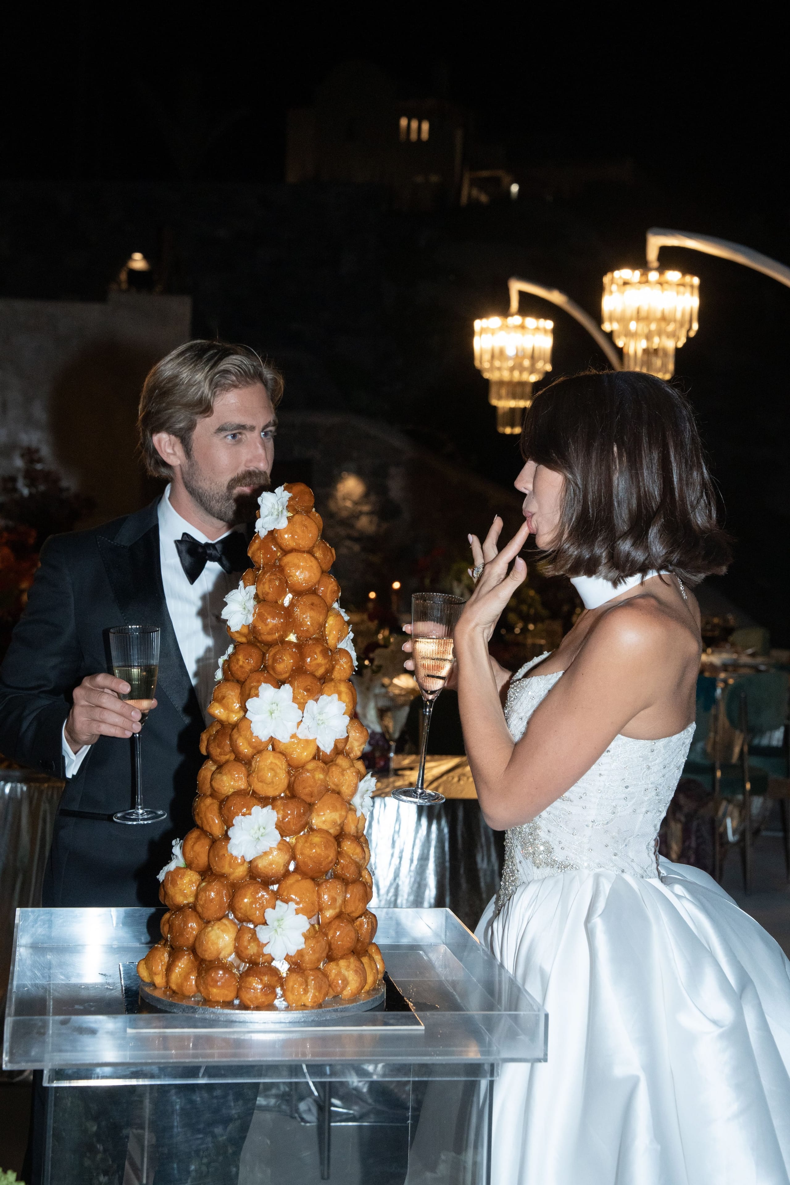 A tall tower of golden choux pastry puffs, decorated with white blossoms, stands on a clear acrylic stand at a wedding reception at Islet Santorini in Greece. Behind it, a man in a black tuxedo and bow tie holds a champagne flute and gazes toward a bride in a strapless white satin gown with a beaded bodice, who lifts a finger to her lips with a flute of champagne in her other hand. Warm glowing chandeliers and a dark evening sky set a romantic, celebratory mood.
