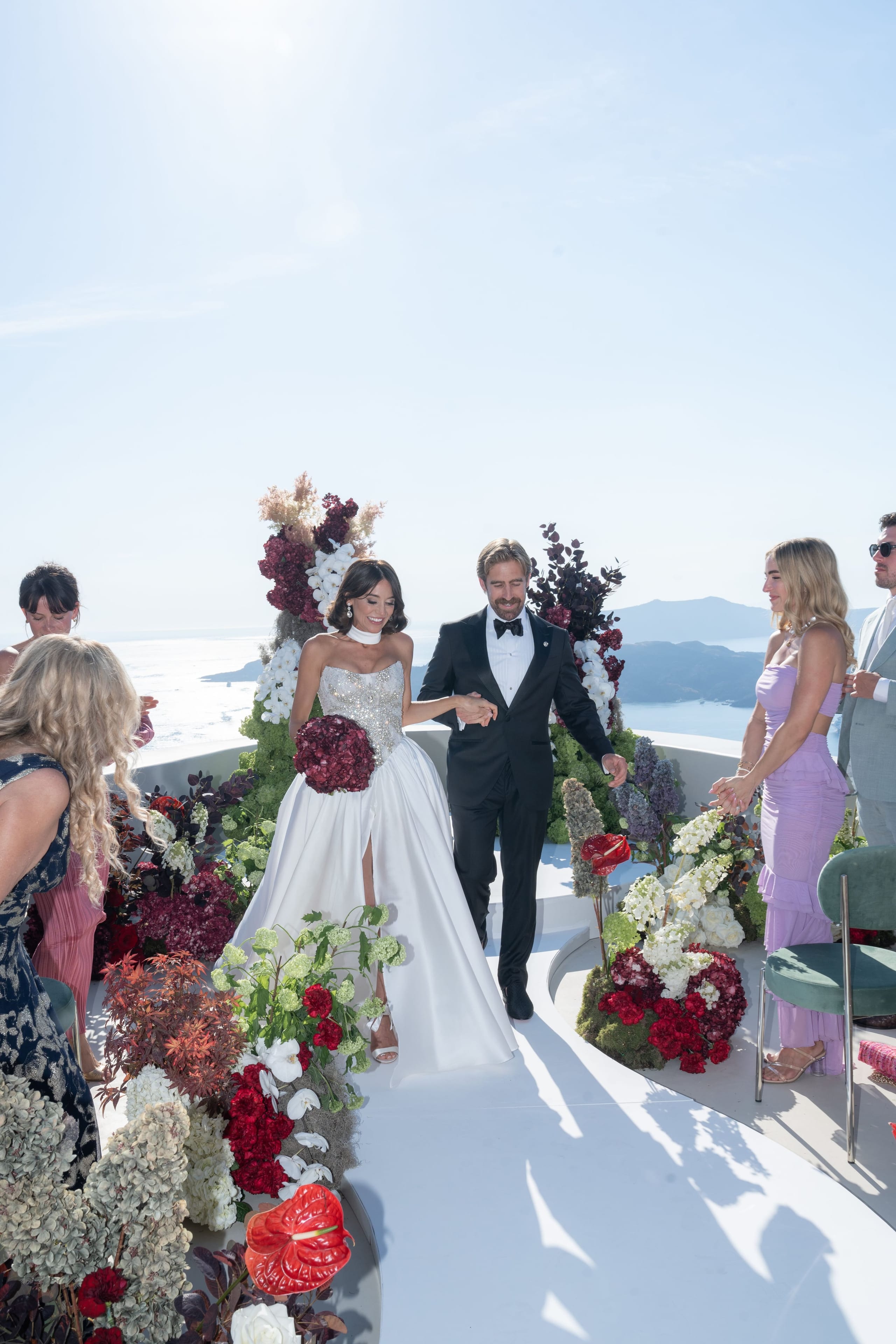A bride in a white satin ball gown with a sparkling embellished bodice and a dark red bouquet walks hand in hand with her groom, who wears a black tuxedo and bow tie, down a white aisle at a wedding in Santorini, Greece. Both sides of the aisle are lined with dramatic floral arrangements of deep burgundy roses, white hydrangeas, white orchids, red anthuriums and dried pampas, with a tall dark-foliaged arch behind them. Guests in lilac, navy and grey stand at the edges, and the bright sky and calm sea of the caldera stretch out behind them.