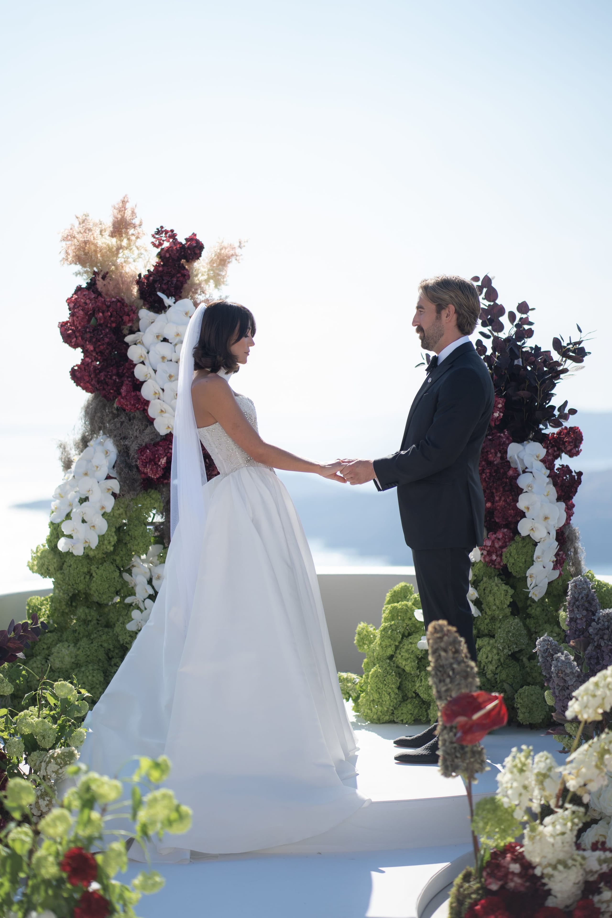 A bride in a strapless white ballgown with a beaded bodice and a long veil holds hands with a groom in a black tuxedo and bow tie, the two facing each other before a wedding arch at Islet Santorini in Greece. The arch is lush with deep burgundy hydrangeas, white phalaenopsis orchids, pale plumes and dark eucalyptus, framed by clusters of green hydrangea, with the soft blue of the Aegean sea and sky behind them.