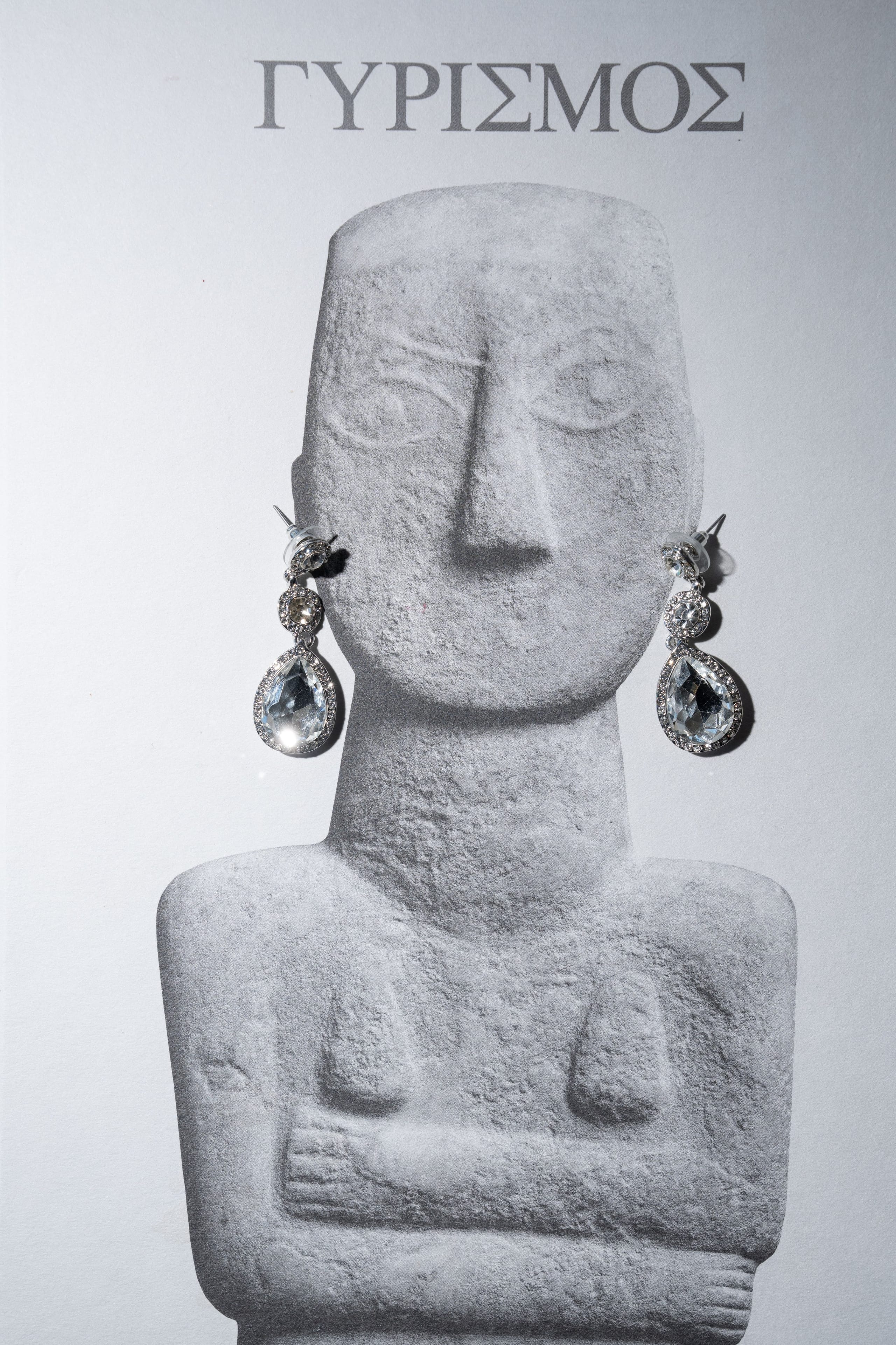 A pair of silver drop earrings, each with a pear-cut clear stone framed in tiny pavé diamonds and a small round stone at the top, hangs from the ears of a grey marble-look figurine, a stylised Cycladic idol with a long face and folded arms. The image is a wedding jewellery display in Santorini, Greece, at Islet, with the word ΓΥΡΙΣΜΟΣ printed in grey above the figure. The background is pale and softly lit, so the sparkle of the stones stands out crisply.