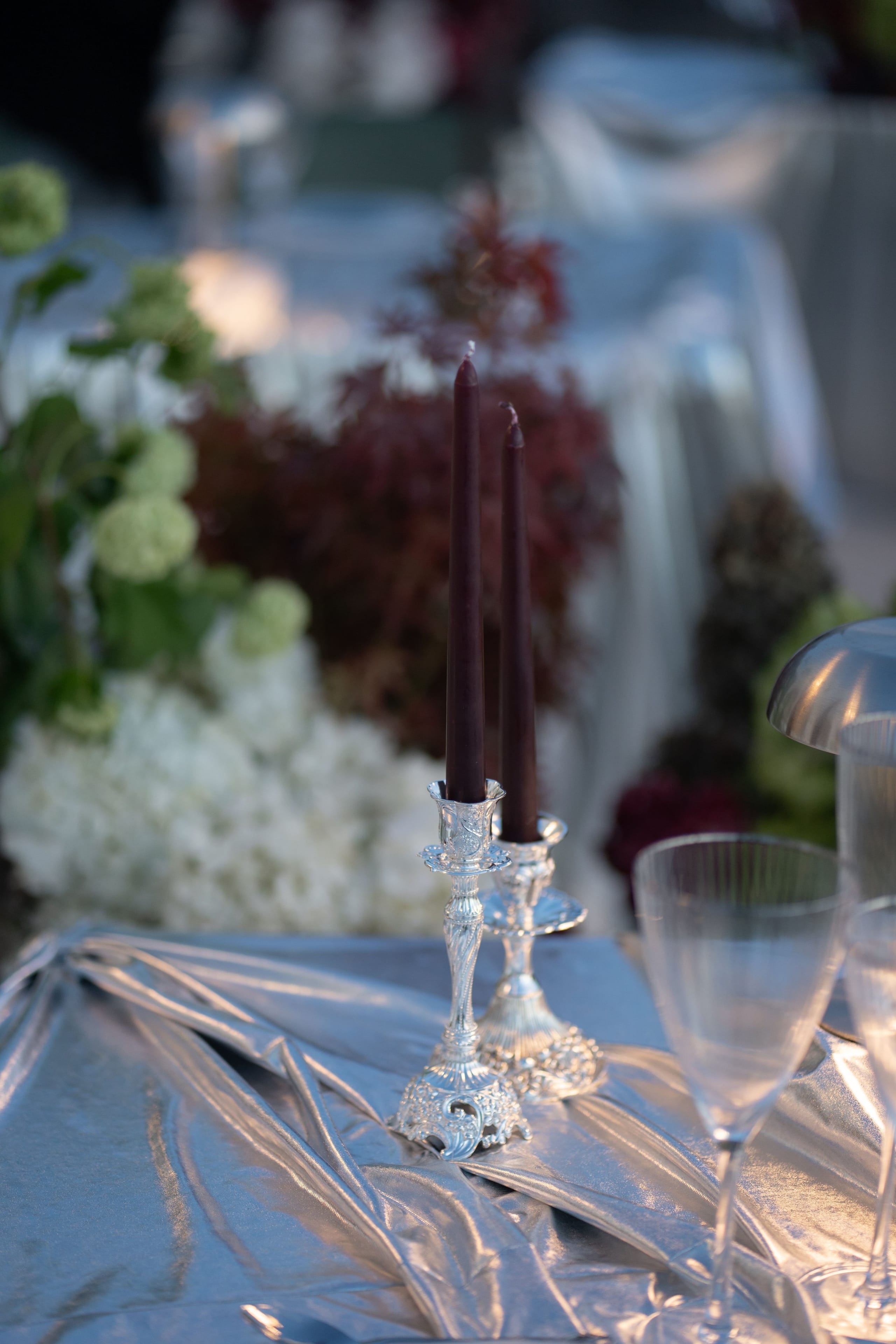 Two tall burgundy taper candles stand in ornate silver candlesticks on a rumpled silver-satin tablecloth, with crystal wine glasses at the right edge. Behind them, soft-focus arrangements of white hydrangeas, green viburnum-style blooms and deep burgundy foliage fill a wedding table at Islet Santorini in Thira, Greece, in warm evening light.