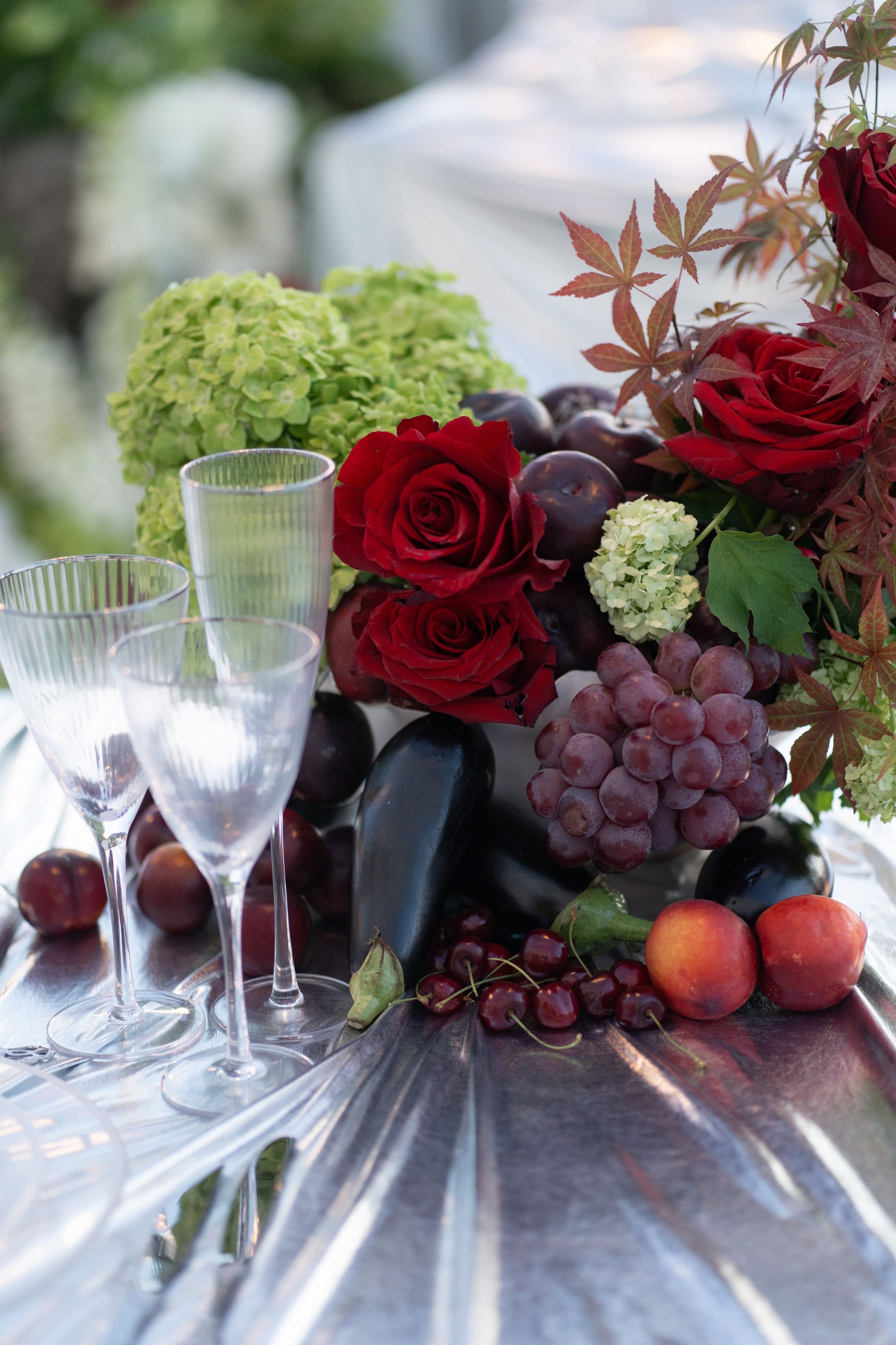 A wedding table centrepiece at Islet Santorini in Thira, Greece, set on a glossy silver tablecloth. Deep red garden roses and sprigs of red Japanese maple rise from a bed of pale green hydrangea, with dark plums, purple grapes, cherries and apricots scattered among them. Two ribbed crystal flutes stand at the left, and the warm evening light gives the whole arrangement a rich, autumnal mood.