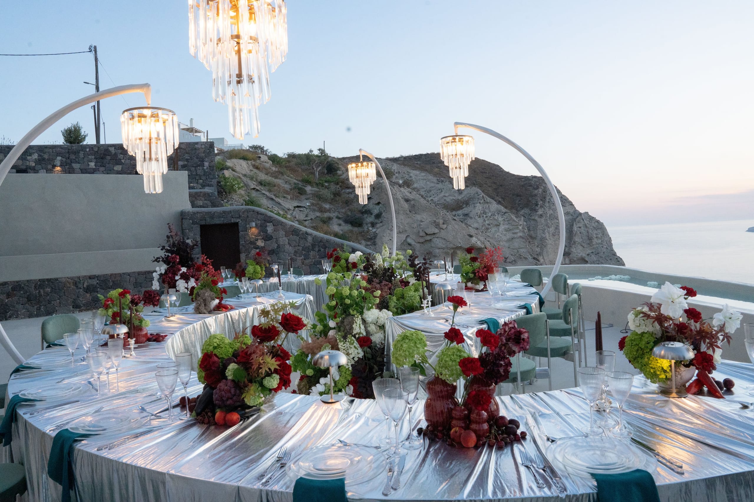 A long curved wedding table on a cliff-top terrace at Islet Santorini in Thira, Greece, dressed in shimmering silver linen with teal napkins and mint-green chairs. Low centrepieces of red garden roses, deep burgundy blooms, green hydrangeas and white flowers sit among small fruits, and crystal chandeliers hang from white arched stands above as the sky fades to dusk over the sea.