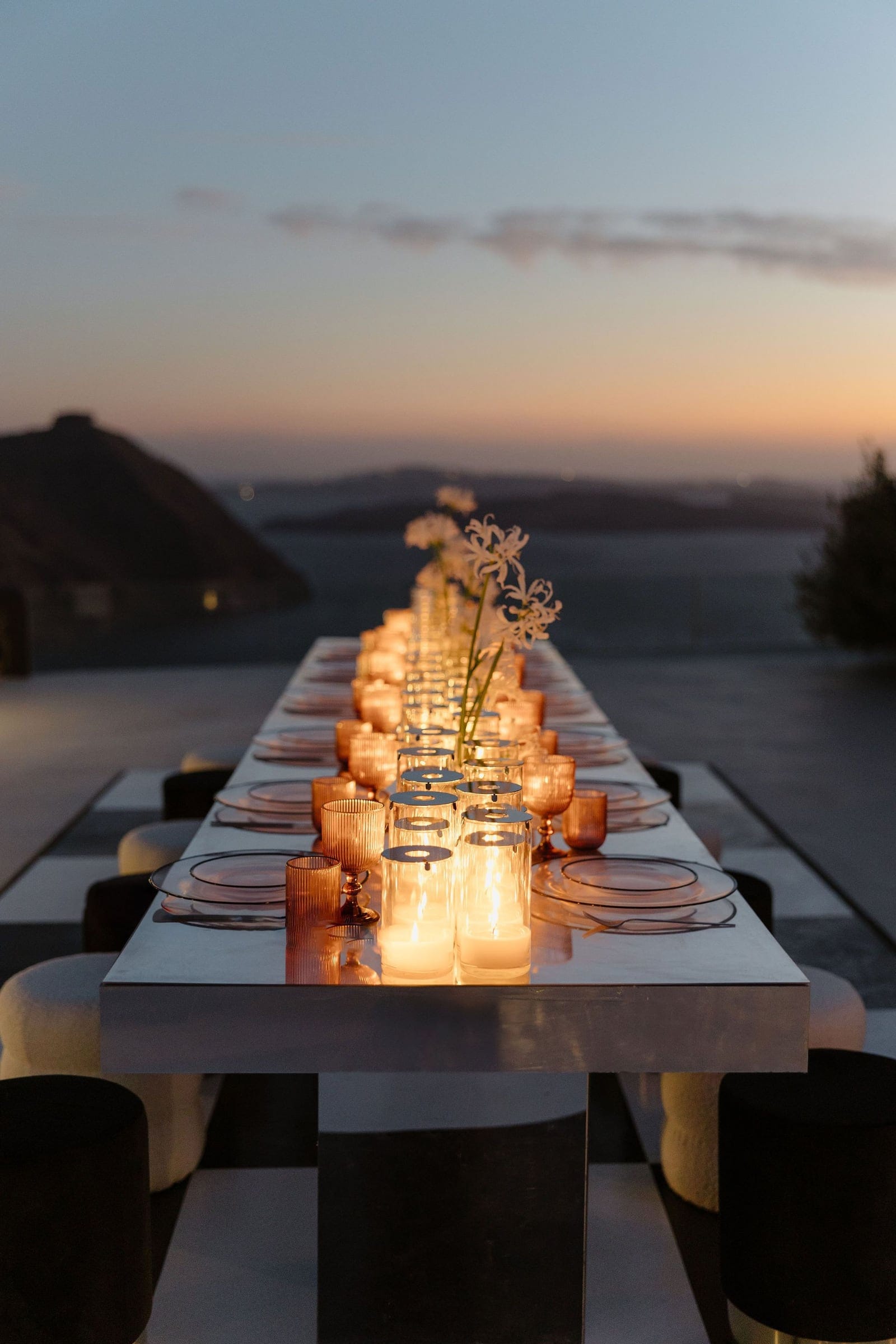 A long white wedding dining table stretches toward the sea at dusk, lined with amber glass goblets, pale plates and tall glass hurricane candles glowing with flame. Along its centre, slender vases hold white, star-shaped flowers, likely spider lilies or similar blooms, with a few stems rising above the rest. Beyond the terrace, the sky fades from soft blue to peach and a hazy dark hill sits on the horizon, a wedding table set at Aenaon Villas in Santorini, Greece.
