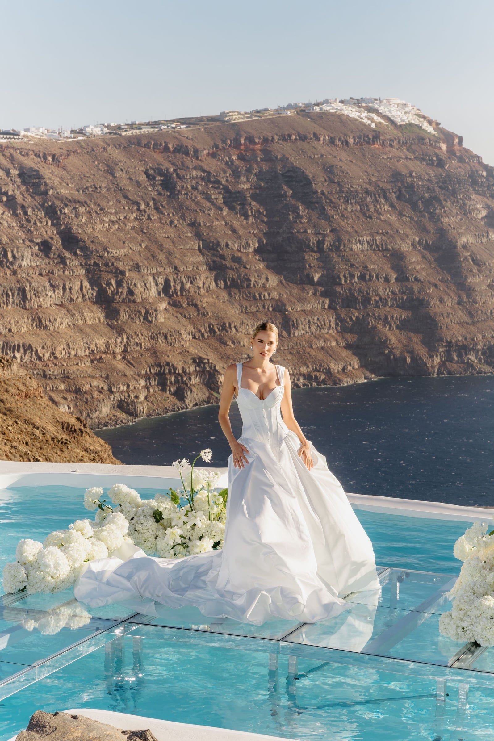 A bride in a white satin gown with a fitted bodice and full skirt stands on a glass platform over a turquoise infinity pool, with white hydrangeas and a trailing white bloom arrangement around her feet. Behind her rises the steep, layered volcanic cliff of the caldera, with white village houses clinging to the ridge above, all set at the wedding flowers venue, Aenaon Villas, on Santorini, Greece.