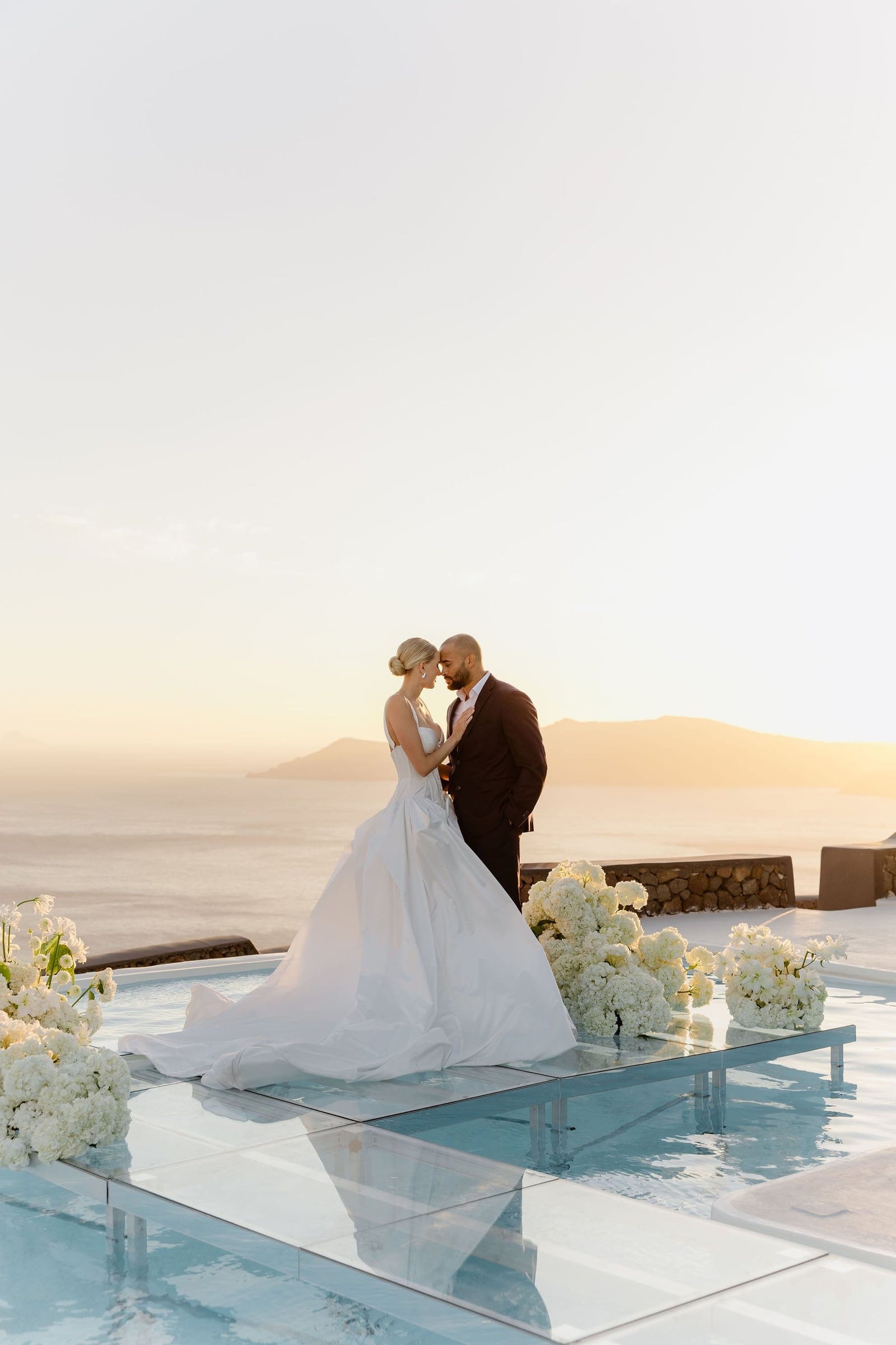 A bride in a voluminous satin ballgown with a long train and a low chignon stands forehead to forehead with her groom in a dark suit, on a glass platform above an infinity pool at sunset. Large white hydrangea and white stock arrangements line the edge of the pool, and the sea and distant hills glow gold behind them at Aenaon Villas, a wedding and event florist setup in Greece.