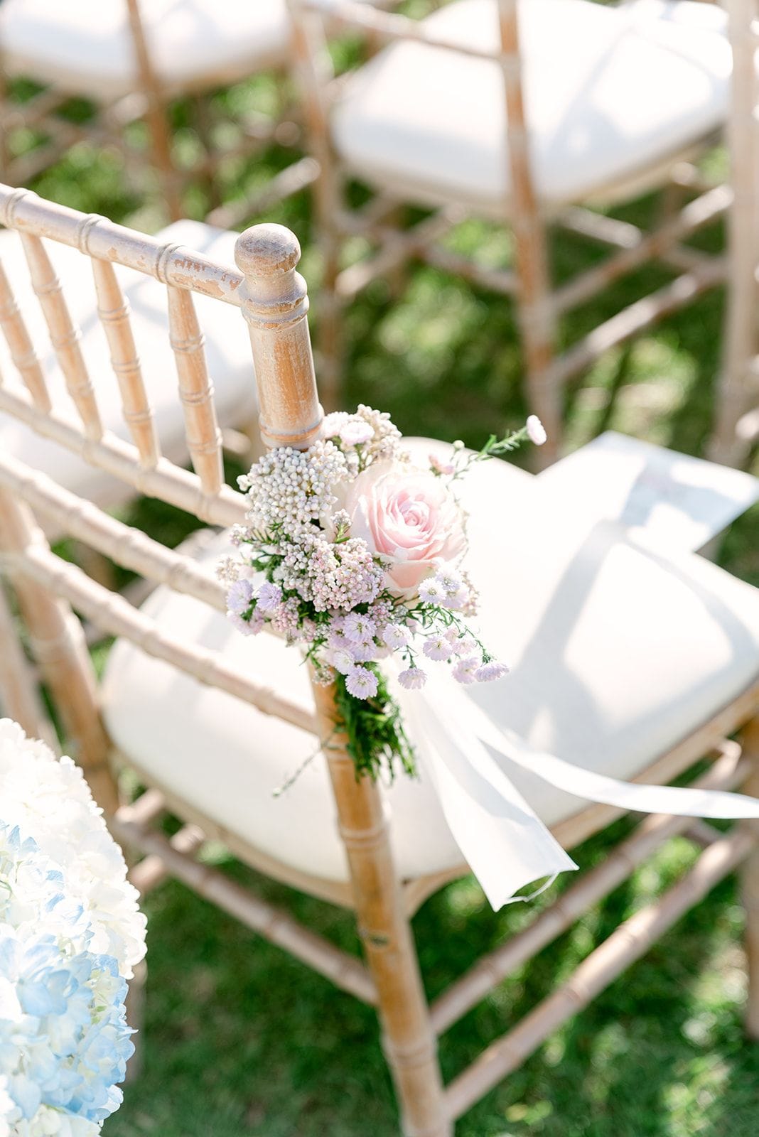 A small posy of a single blush-pink garden rose, tucked among tiny white and lilac blooms and feathery sprigs of gypsophila, is tied to the back of a pale wooden chiavari chair with a long white ribbon trailing over the cushion. Rows of matching chairs stretch away across green lawn in soft, dappled sunlight, with a pale blue and white hydrangea just visible at the lower left. These are wedding ceremony flowers at Κτήμα Χατζή in Κερατέα, Athens, Greece.