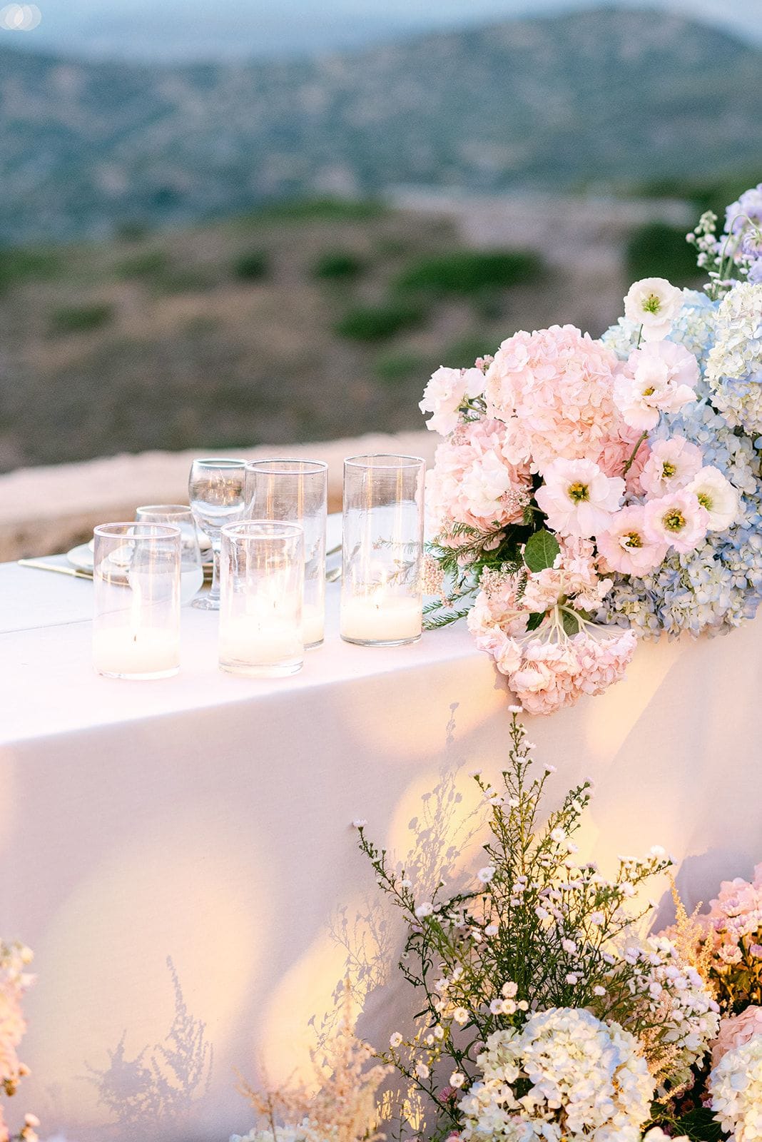 A long white-draped wedding table at dusk, its edge softened by a cascade of pale pink and blue hydrangeas mixed with white and blush anemone-style blooms, with a tumble of white wildflowers and feathery foliage spilling along the floor below. Three clear glass cylinders holding lit pillar candles and a stemmed wine glass sit on the cloth against a backdrop of hazy green hills. This is a wedding table setting at Κτήμα Χατζή in Kerateas, near Athens, Greece.