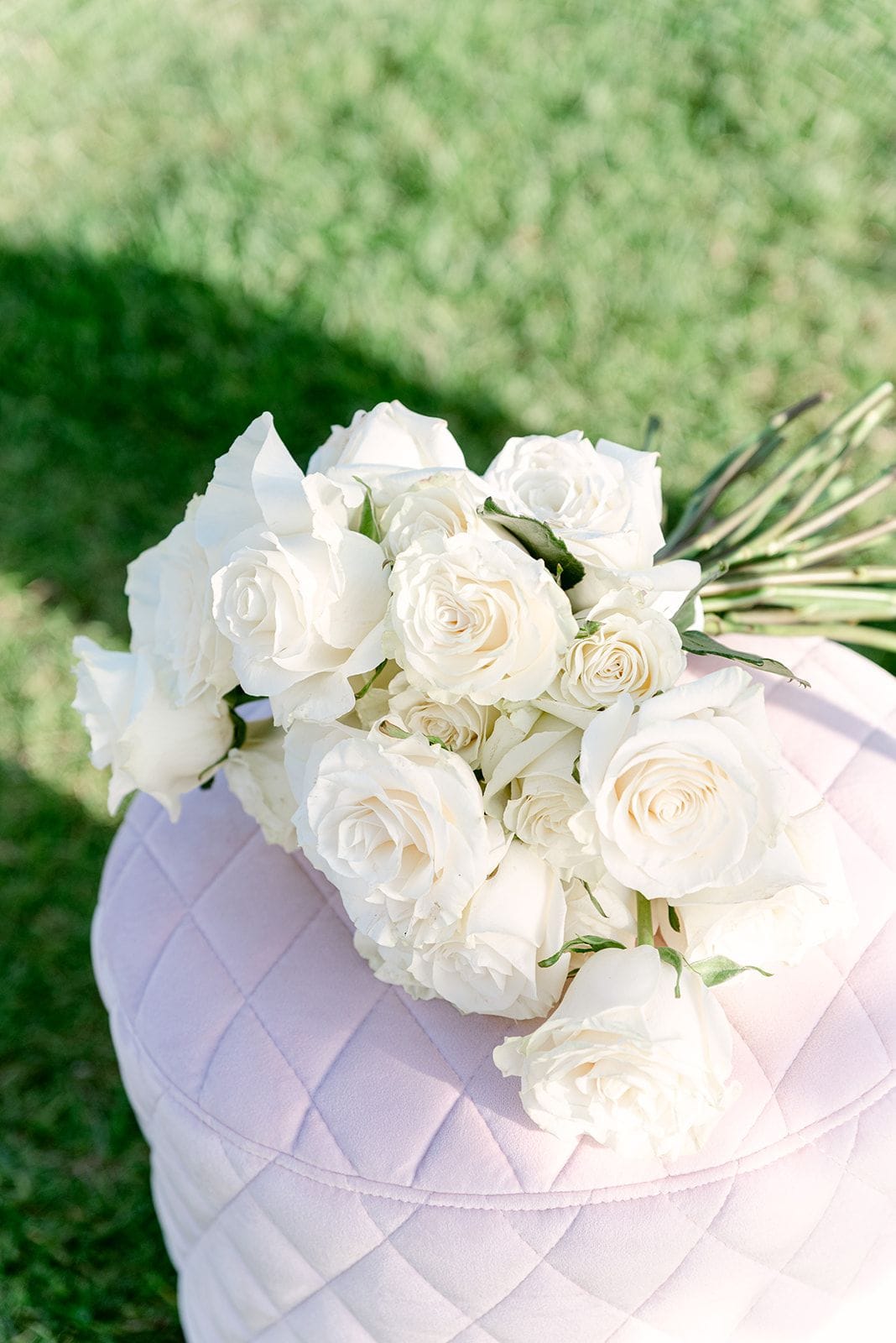 A bridal bouquet of creamy white garden roses, their petals soft and full, with a few green leaves and long stems showing at the right, rests on a pale pink quilted ottoman set on lawn. Sunlight falls across the grass behind it, giving the scene a calm, airy feel. This is a wedding bouquet by Manfler, photographed at Κτήμα Χατζή in Kerateas, Athens, Greece.