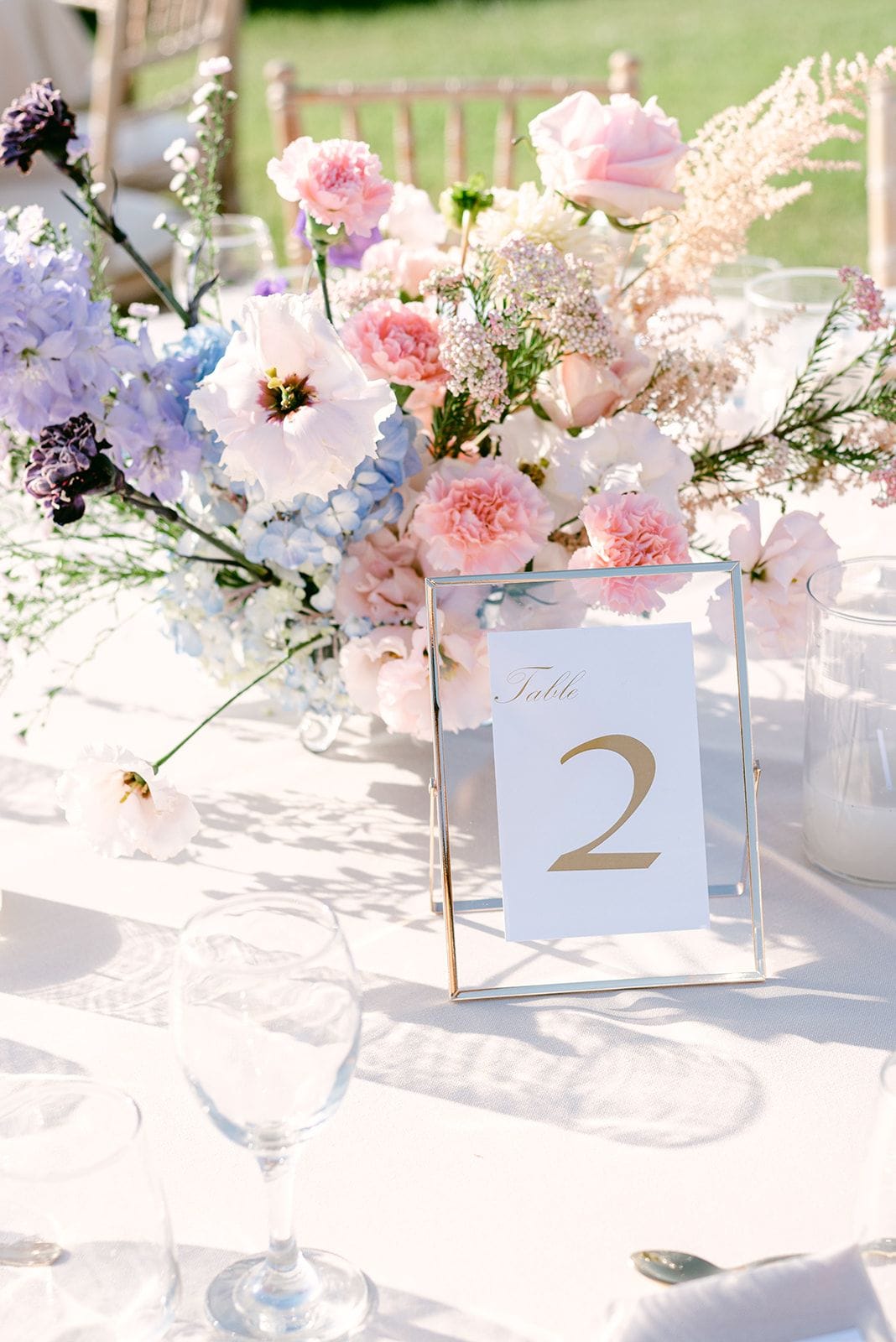 A low centrepiece of pastel blooms sits on a white linen table: pale pink carnations and garden roses, lavender stock, blue hydrangea, a white lisianthus, and feathery astilbe and yarrow. Beside it, a slim gold-framed glass stand holds a white card reading "Table 2" in gold script, and a wine glass catches soft shadows. This is a wedding table flower arrangement at Κτήμα Χατζή in Kerateas, Athens, Greece, with a sunny lawn and empty chairs blurred behind.
