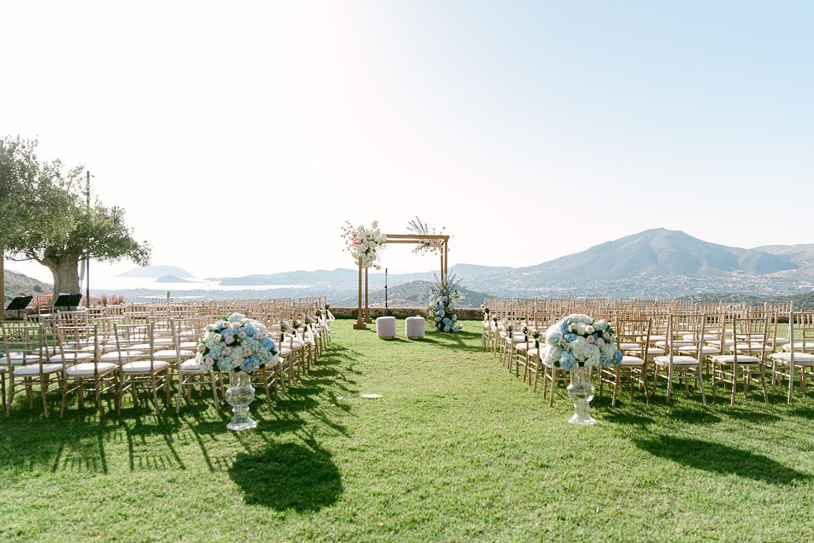 A wedding ceremony aisle on a grassy lawn at Κτήμα Χατζή in Κερατέα, Athens, Greece, lined with rows of gold chiavari chairs with white cushions. Two tall glass urns of white hydrangeas, blue hydrangeas and pale pink roses mark the aisle, and a slim gold wooden arch at the far end is softly dressed with white and blush blooms. Behind it, a wide sea view and hazy mountains stretch under a pale summer sky, with an old olive tree shading the left side.