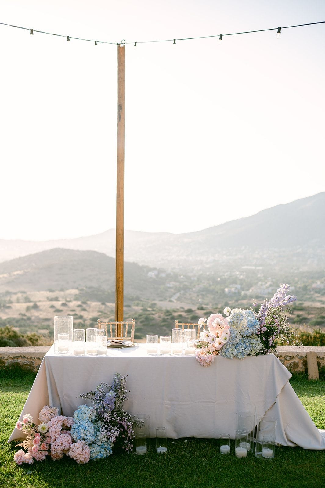 A long white-draped table sits on a lawn at a hilltop venue in Kerateas, near Athens, Greece, set for a wedding under a string of bulb lights strung from a tall wooden pole. Pastel arrangements of blue and pink hydrangeas, pale pink garden roses and lavender delphinium spill from each end of the table and down to the grass, with clusters of glass candle holders along the front. Soft, hazy light falls over the rolling hills and village below, giving the scene a calm, golden-hour mood.