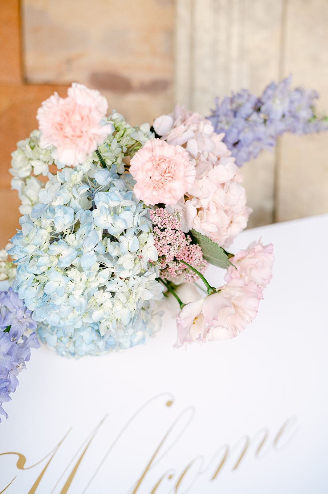 A pastel arrangement of pale blue hydrangeas, pink carnations, blush lisianthus, and lilac delphinium sits on a white table at a wedding at Κτήμα Χατζή in Κερατέα, near Athens, Greece. Clusters of tiny pink yarrow and soft green leaves fill the gaps, and gold script lettering reading "Welcome" runs along the bottom edge of the table.