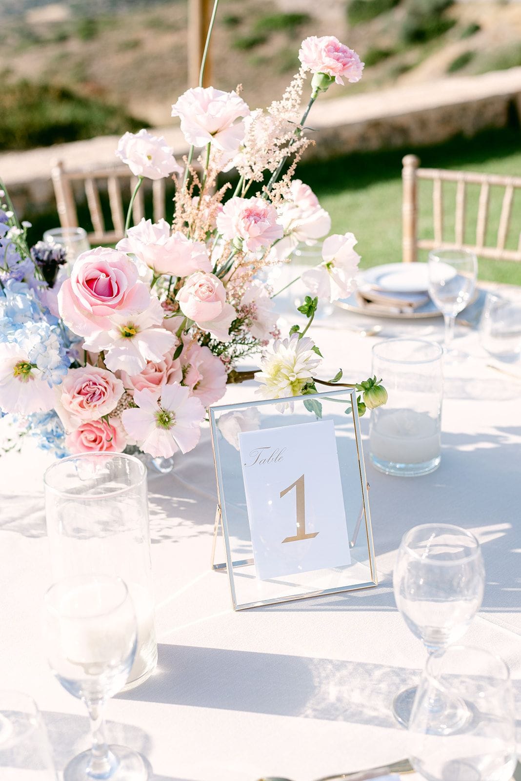 A wedding table centrepiece of soft pink garden roses, pale pink carnations, white anemone-like blooms and feathery astilbe, with trailing blue delphinium at one side, set on a white linen tablecloth. In front, a slim gold-framed glass stand holds a card reading "Table 1" in gold script. This is a wedding in Kτήμα Χατζή in Keratea, Athens, Greece, on a sunlit terrace with dry hillside behind.