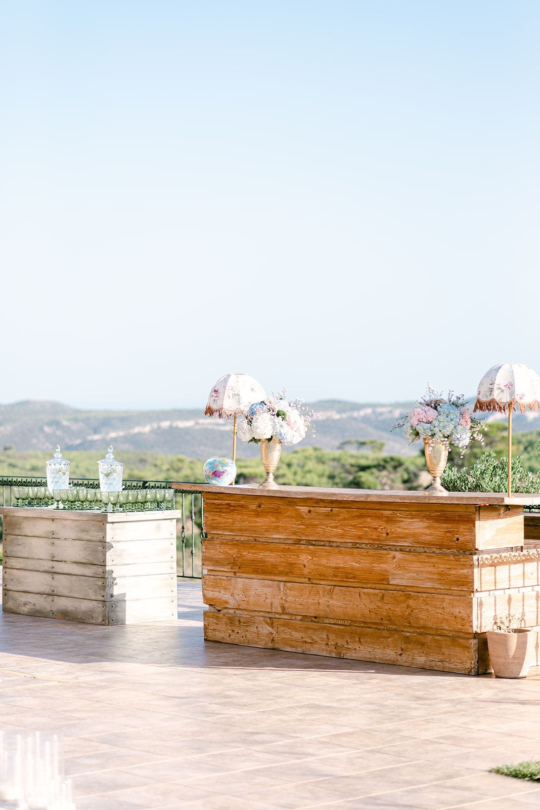 A rustic wooden bar stands on a stone terrace, decorated with two gold-footed vases of white, pale blue and blush-pink hydrangeas and baby's breath, each beneath a small fringed parasol. To the left, a weathered whitewashed crate holds glass canisters and rows of stemmed glasses. Beyond the terrace, pine-covered hills stretch under a pale sky, marking this wedding flower display at Κτήμα Χατζή in Κερατέα, Athens, Greece.