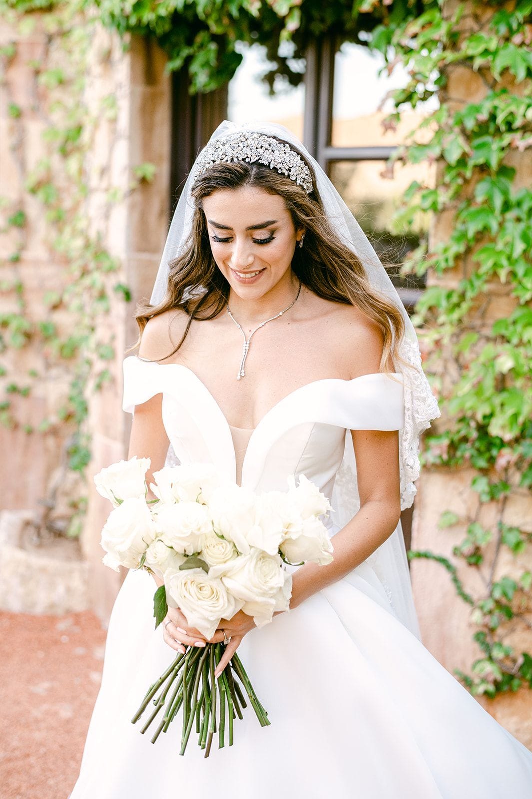 A bride in an off-the-shoulder white gown and a sparkling jewelled headband with a lace veil stands smiling softly, eyes lowered toward her bridal bouquet of creamy white garden roses with long green stems. Behind her, ivy climbs a warm stone wall beside a window, at Κτήμα Χατζή in Κερατέα, Athens, Greece.