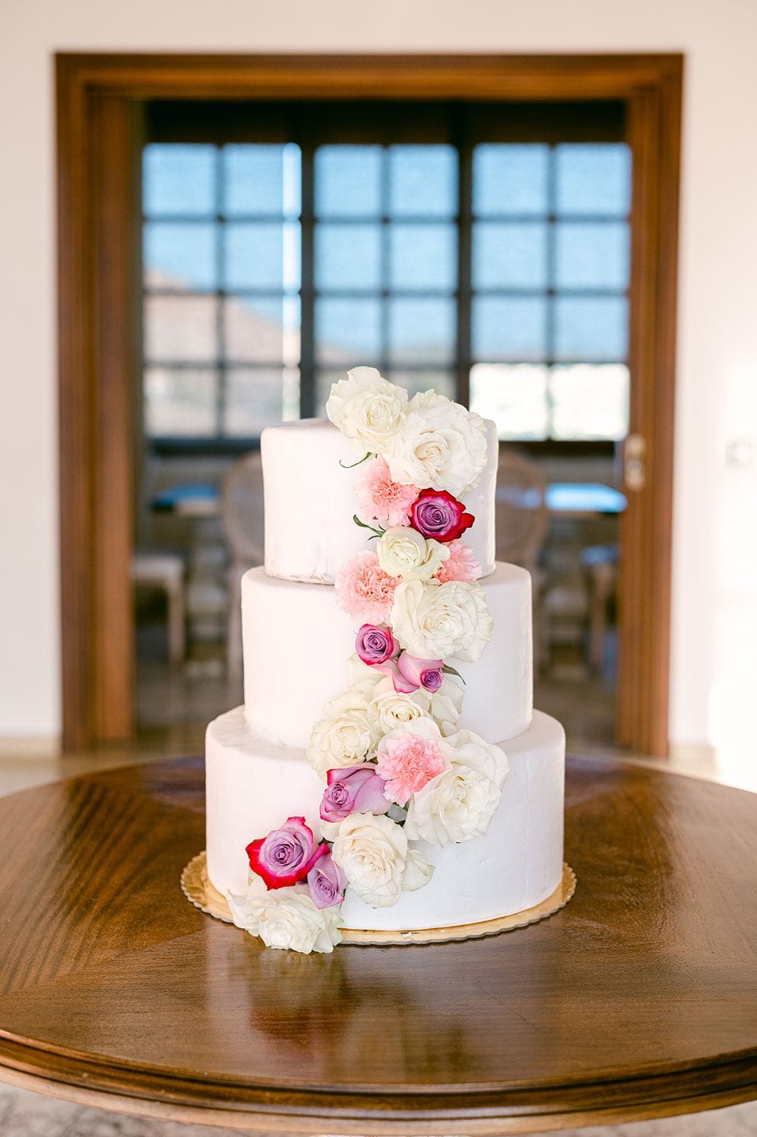 A three-tier white buttercream wedding cake on a round wooden table, decorated with a cascade of white garden roses, pink carnations, and magenta and lavender roses winding down one side. Tall windows and a wooden doorframe glow with soft daylight behind it at Κτήμα Χατζή in Κερατέα, Athens, Greece.