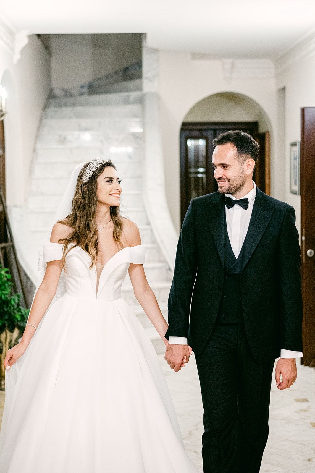 A bride and groom walk hand in hand through a grand interior at Ktima Xatzi in Greece, smiling at each other. The bride wears an off-the-shoulder white ballgown with a deep V neckline and a sparkling crystal headpiece with a long veil, while the groom wears a black tuxedo with a bow tie. Behind them a wide white marble staircase curves upward, framed by an arched wooden doorway, with a trailing green plant at the left edge. The soft, warm light gives the scene a calm, elegant mood.