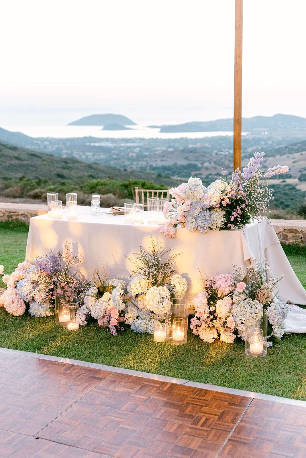 A long white-draped head table set on a grassy hilltop terrace at Κτήμα Χατζή in Κερατέα, Athens, Greece, dressed with wedding flowers in soft pink, lilac and cream. Clusters of white and pale blue hydrangeas, pink roses and sprays of delphinium and baby's breath spill from low arrangements along its base and from a tall bouquet at one end, with lit candles in glass hurricanes glowing among them. Beyond the terrace, the land falls away to a hazy sea and distant islands in the warm, pastel light of early evening.