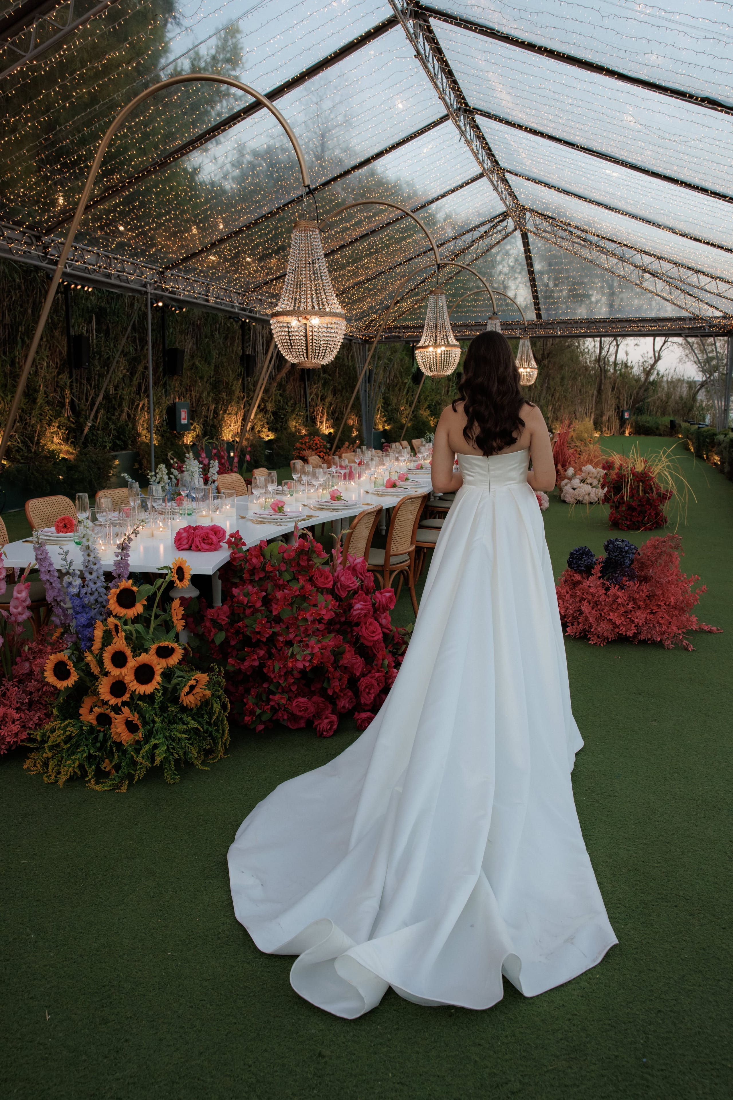 Wedding table flowers at Island Club & Restaurant in Vari, Athens, Greece: a bride in a strapless white satin gown with a long sweeping train walks away from the camera, toward a long white table set for dinner. Beneath a clear canopy strung with fairy lights and hung with crystal chandeliers, the floor is lined with lush clusters of sunflowers, deep red bougainvillea, pink roses and dark blue hydrangeas on green lawn.