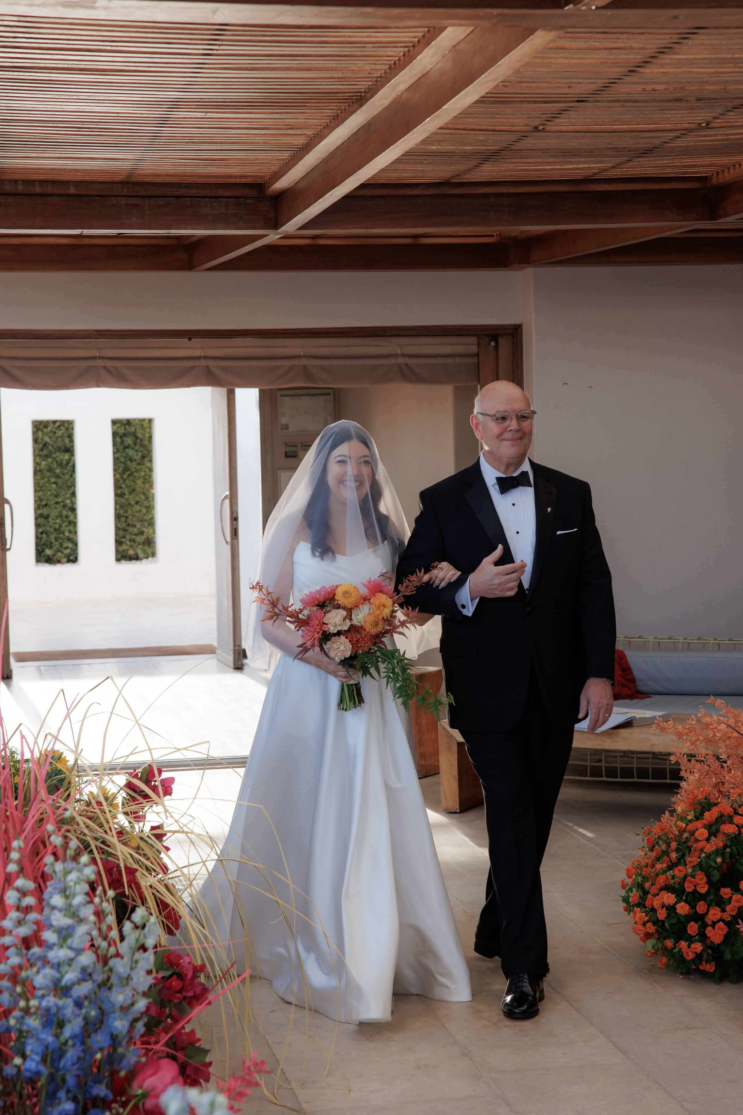 A bride in a satin ballgown and sheer veil walks down an aisle beneath a wooden slatted ceiling, holding a bouquet of orange and coral dahlias, peach roses and warm autumn foliage, her arm linked with an older man in a black tuxedo and bow tie. Along the aisle stand tall spires of blue delphinium, red celosia and orange flowers in bold arrangements, all part of the wedding ceremony flowers at Island Club & Restaurant in Vari, Athens, Greece.